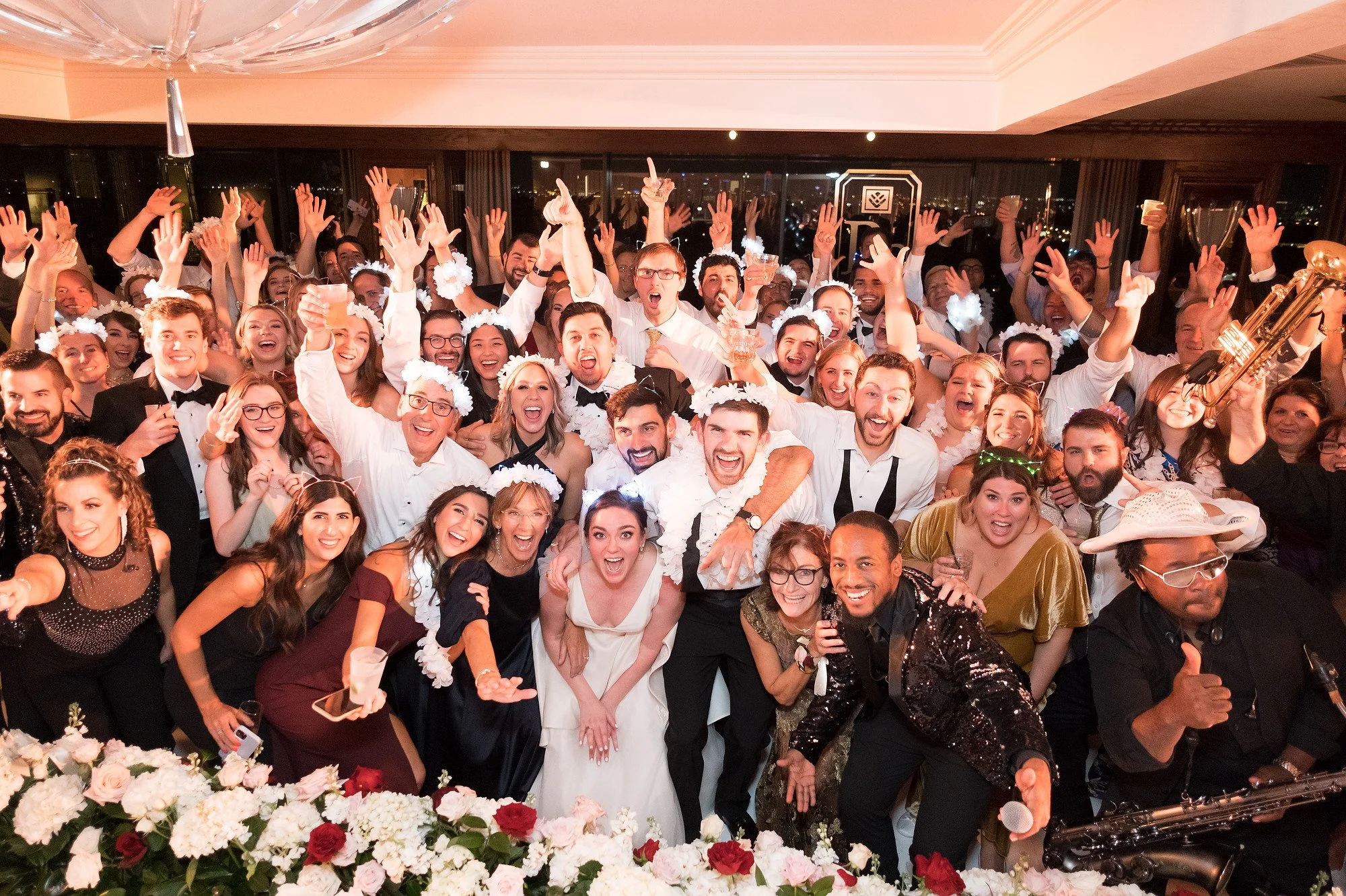 A large group of people at a celebration or wedding party, smiling, cheering, and raising their hands in the air inside a decorated venue with floral arrangements in the foreground.