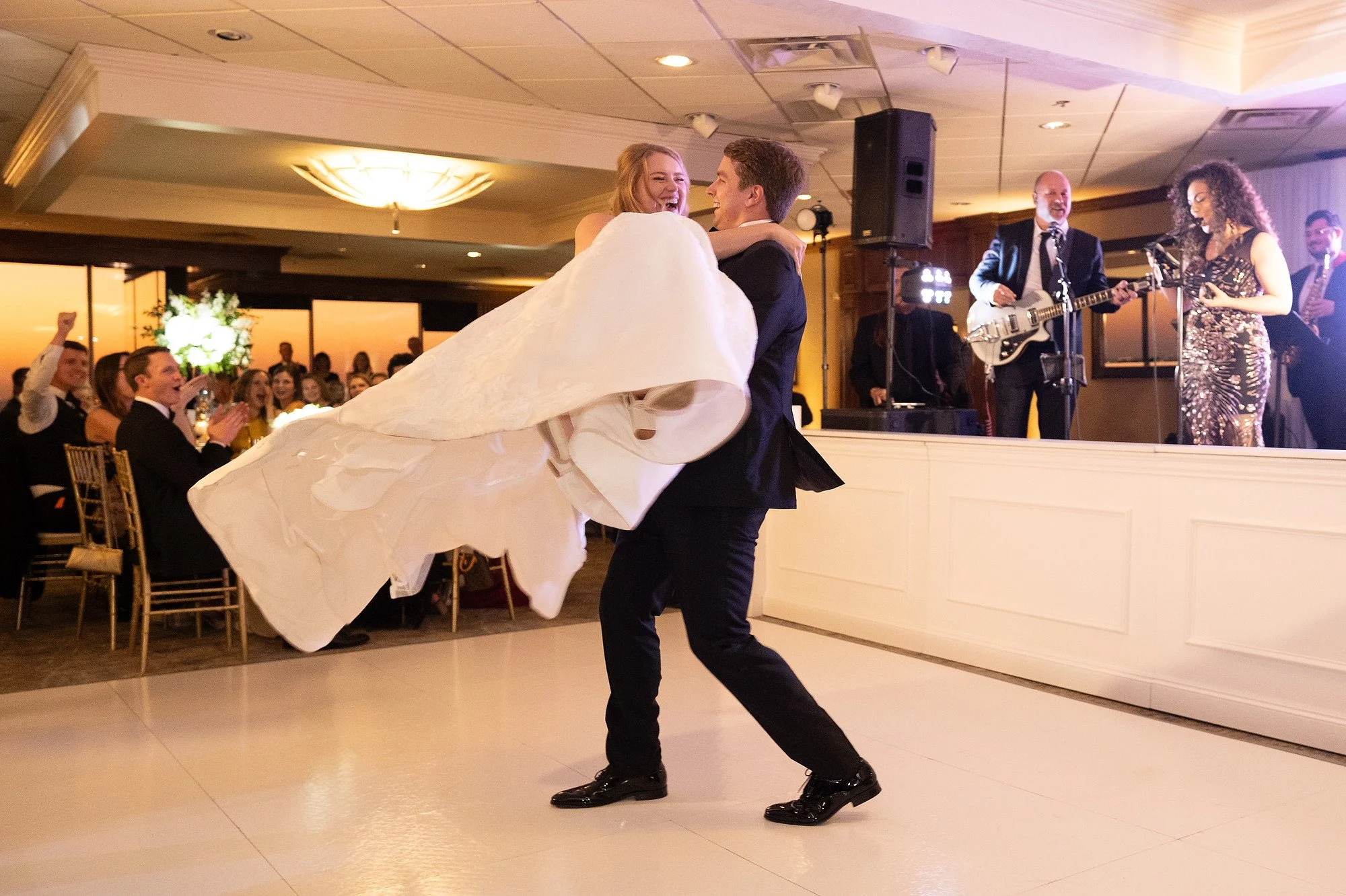 Groom dancing with bride in wedding gown at reception while band plays in background.