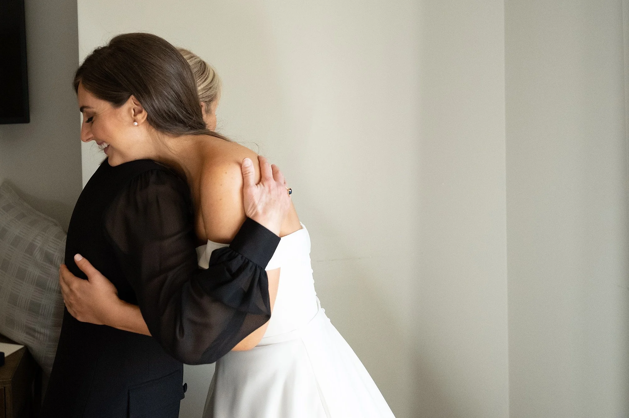 Two women hugging, one in a black dress and the other in a white wedding gown, in a warmly lit room.