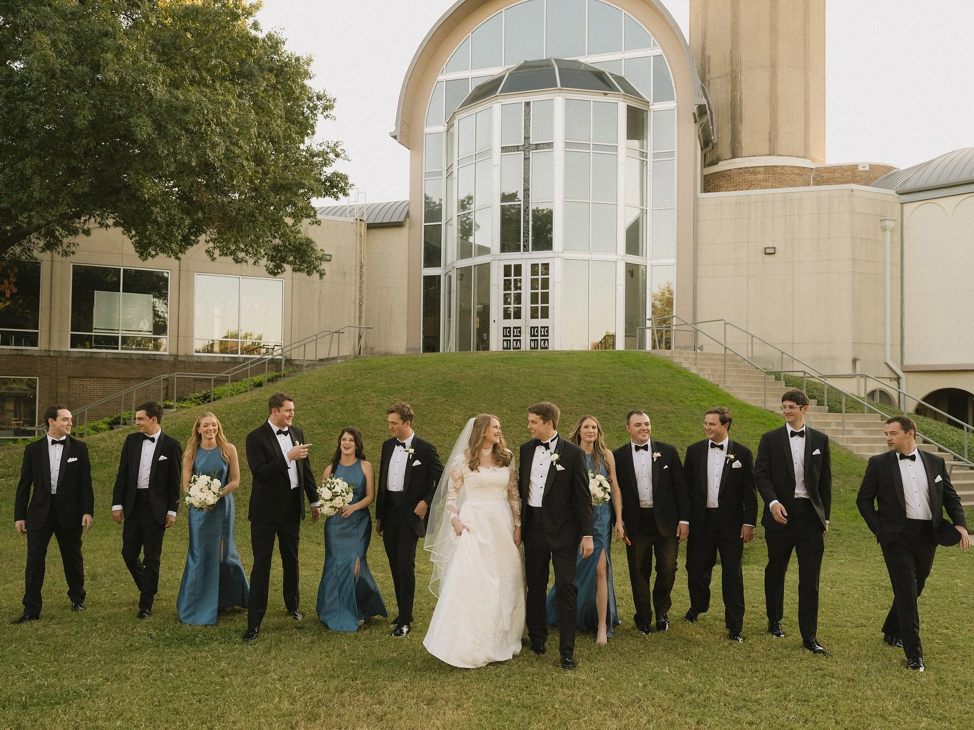 Wedding party consisting of bridesmaids in teal dresses and groomsmen in black tuxedos walking together outside on a grassy area near a modern church building.