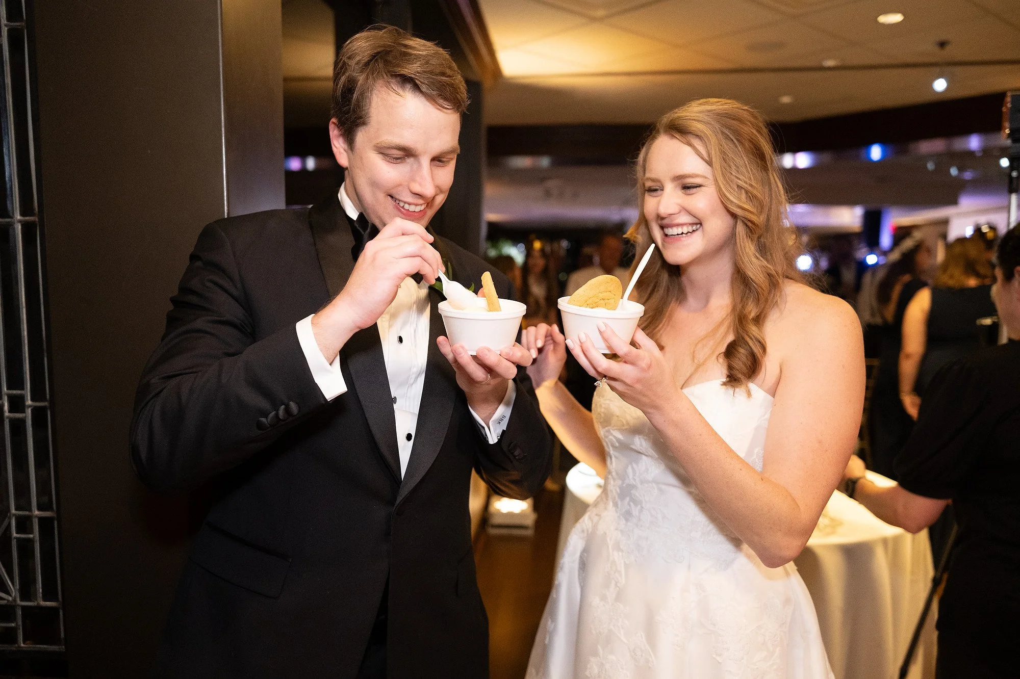 A bride and groom in wedding attire sharing and enjoying cups of ice cream at their wedding reception.