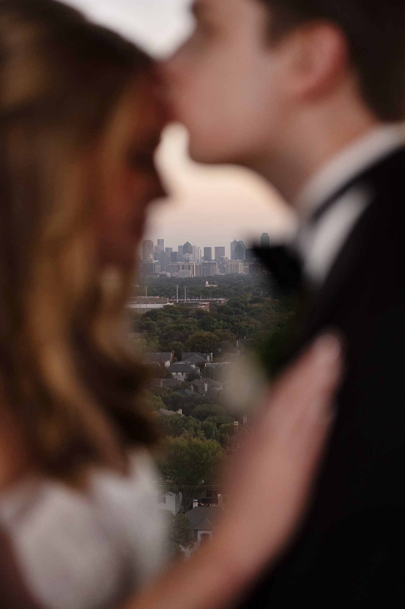 A blurry close-up of a couple in formal attire sharing an intimate moment, with a city skyline in the background.