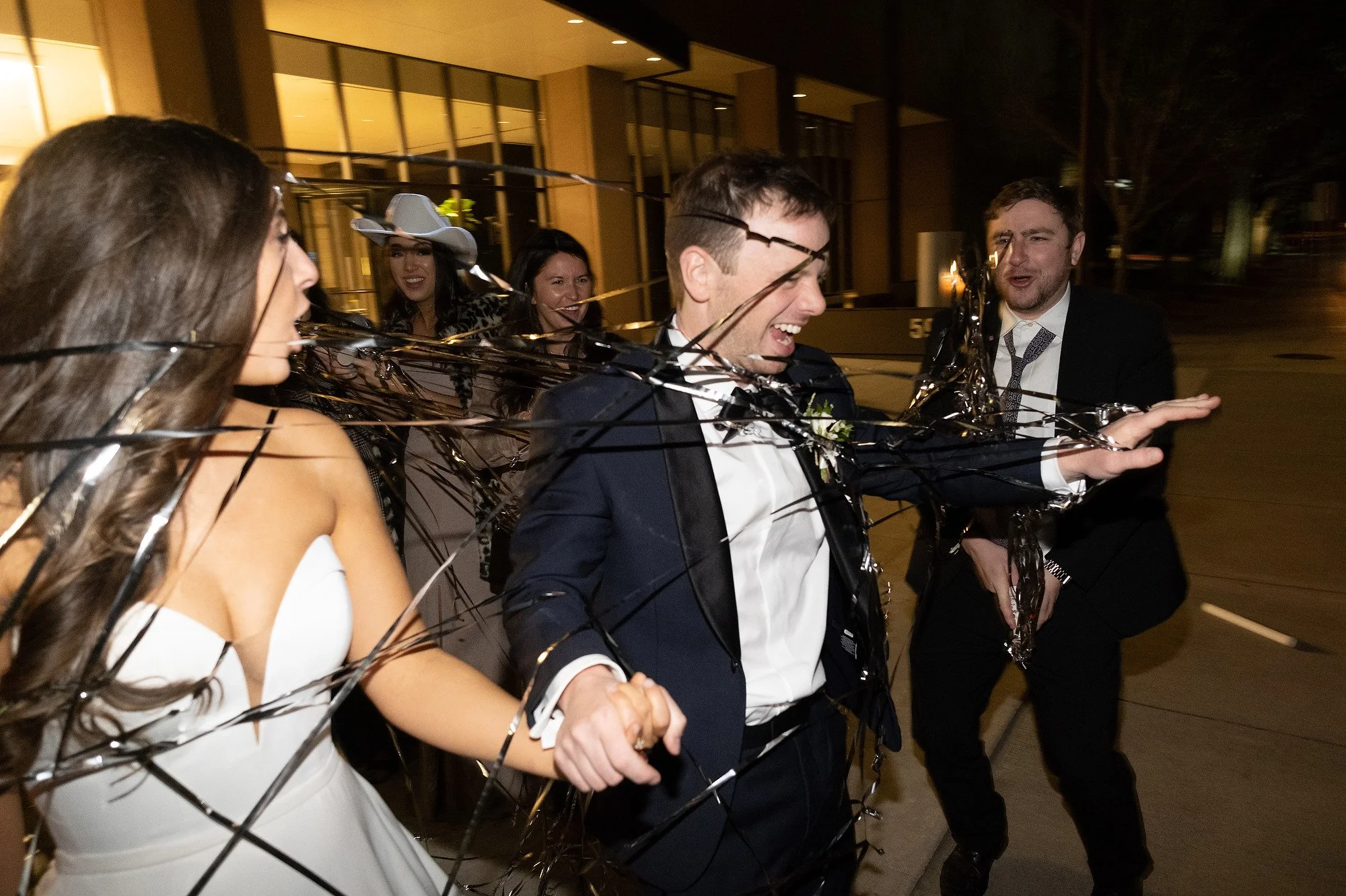 A group of people celebrate at night with a wedding party, some wrapped in black tinsel, smiling and laughing outside a building.