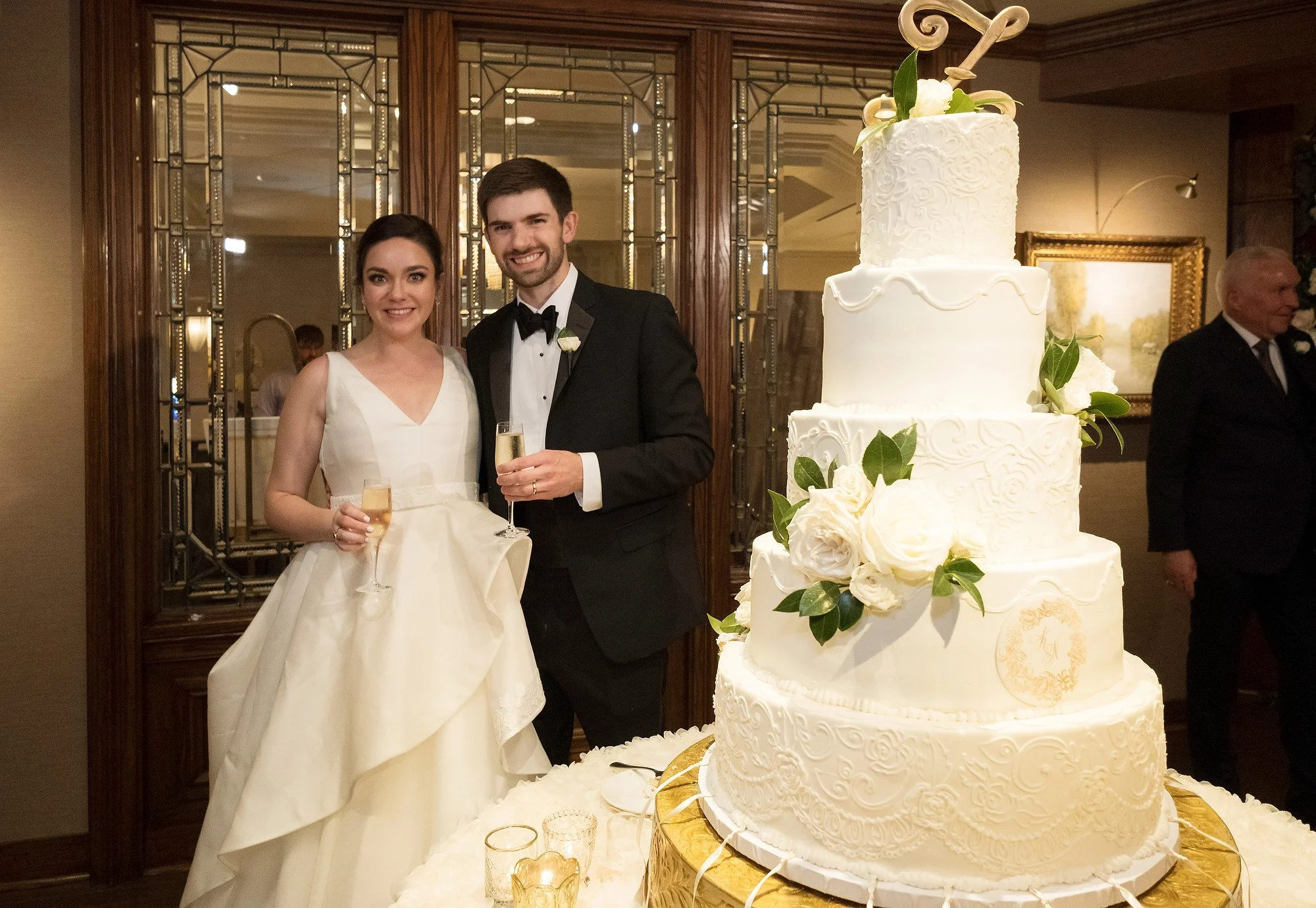Newlywed couple in wedding attire holding champagne glasses, standing next to a large white wedding cake decorated with white roses and green leaves at reception.