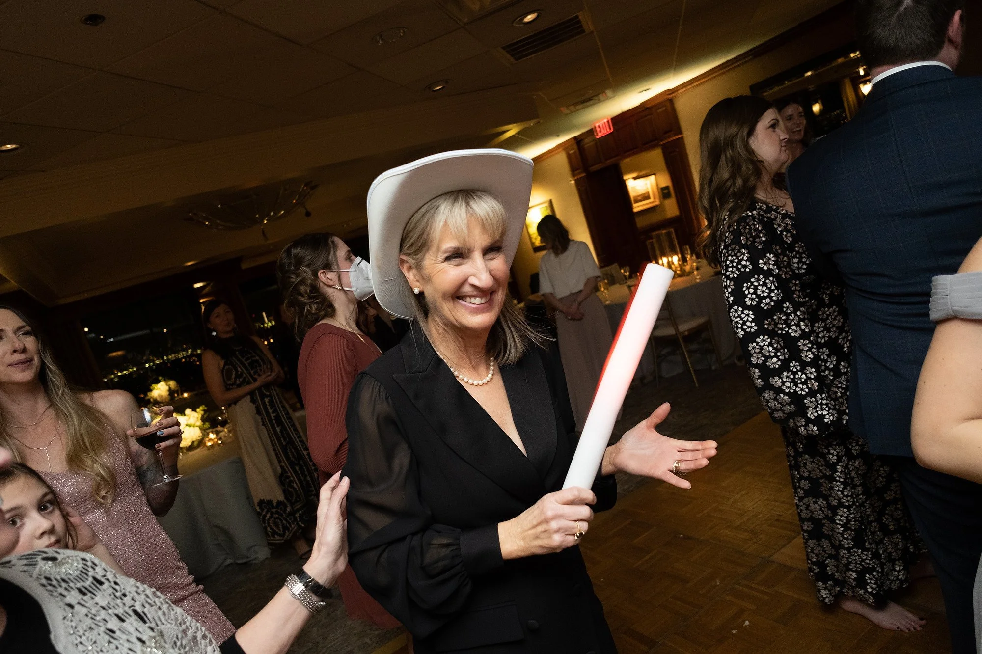 A woman wearing a large white hat, black outfit, and pearl necklace is smiling and holding a white glow stick at a party or celebration, surrounded by other people in a warmly lit room.