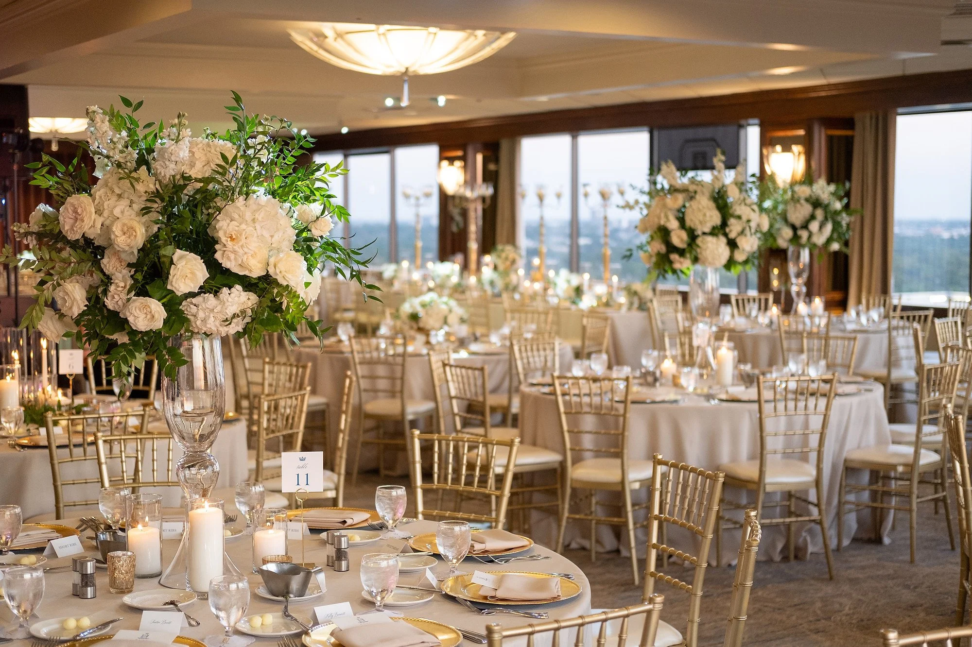 Elegant banquet hall decorated with tall floral centerpieces of white flowers and candles on tables with gold Chiavari chairs and large windows overlooking the city.
