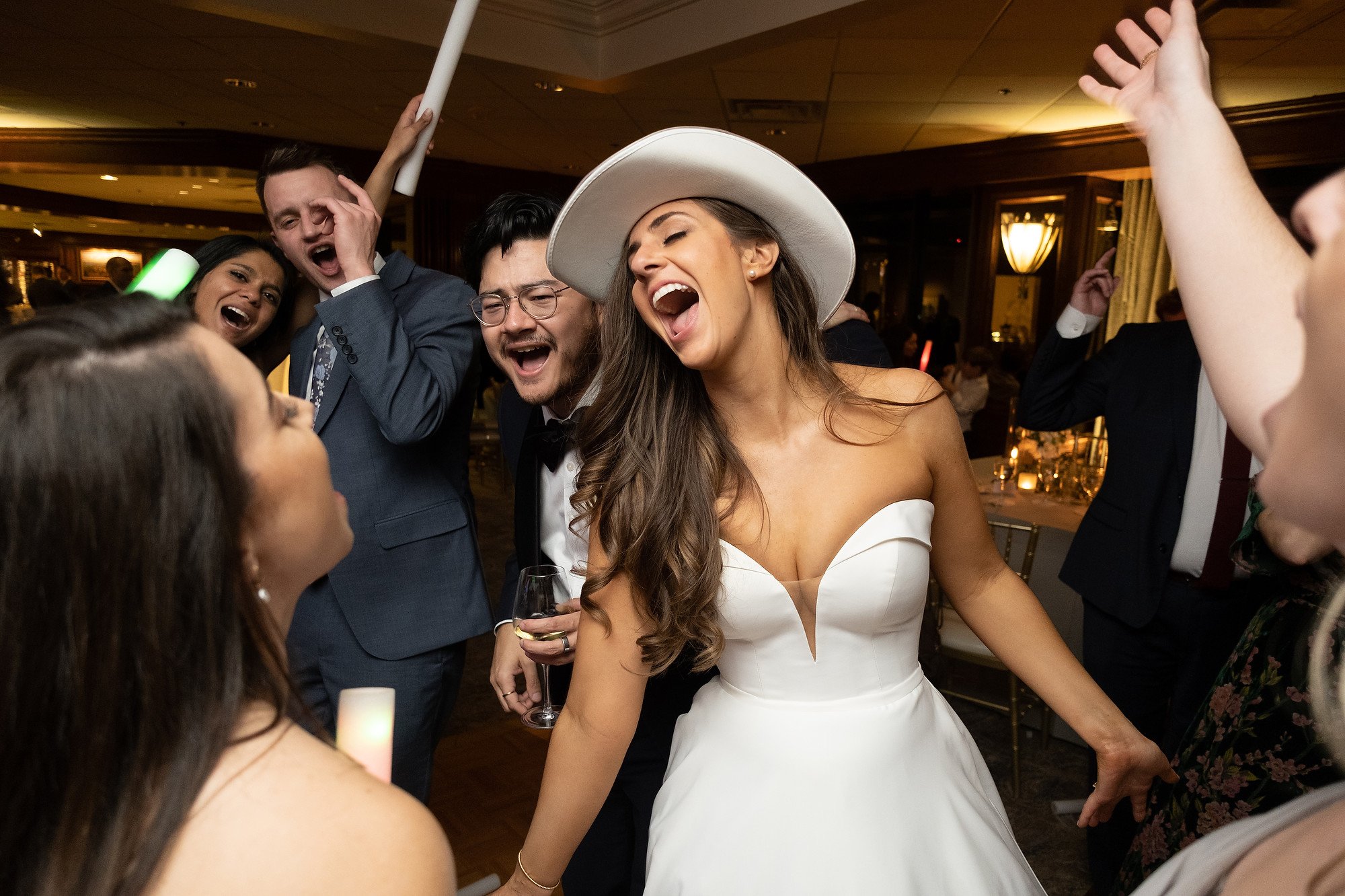 Bride in a white strapless wedding dress and wide-brimmed hat dancing and laughing with friends at a wedding reception, surrounded by guests in formal attire.