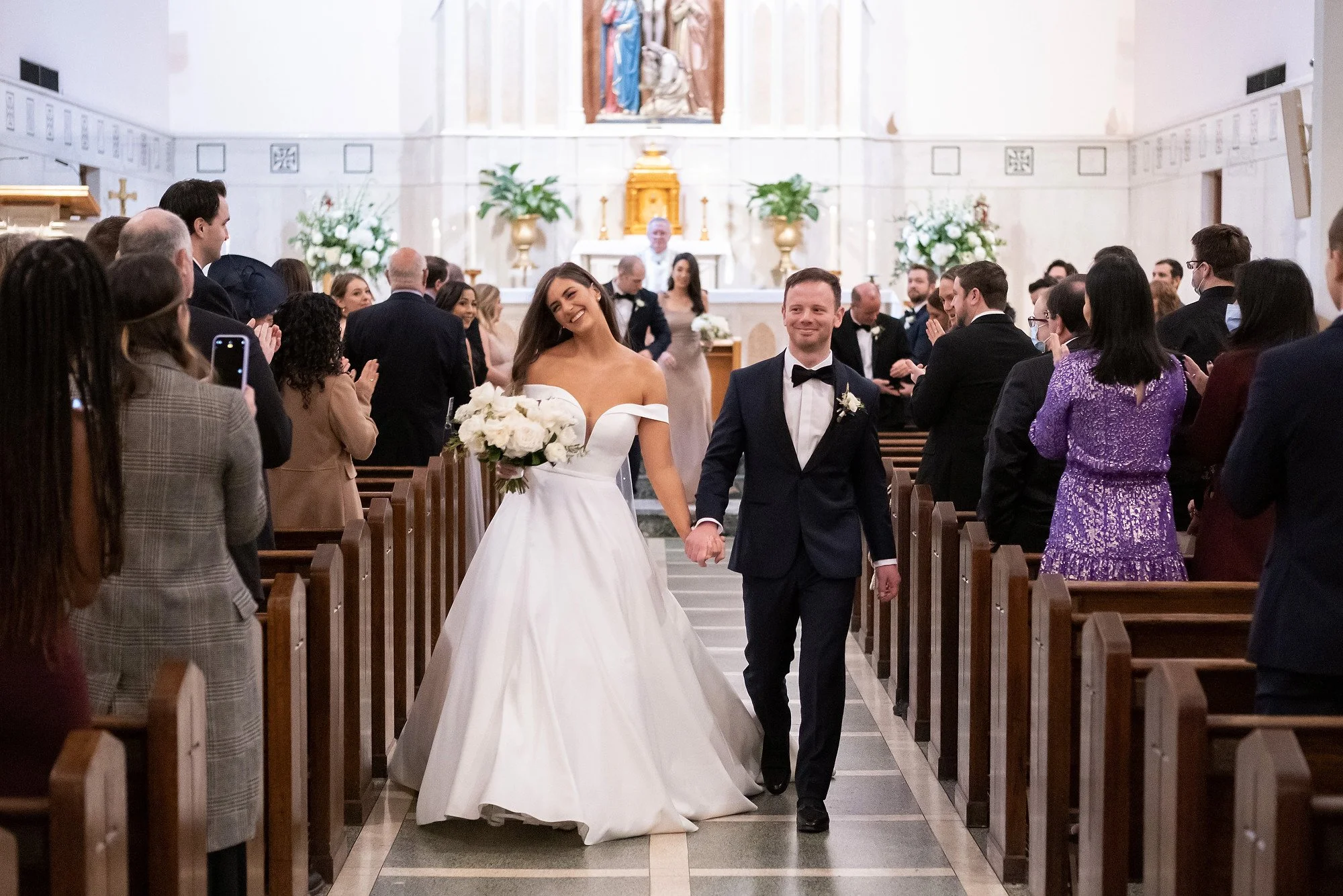 a bride and groom walk down the aisle of a church after their wedding, holding hands and smiling, with wedding guests standing and applauding on both sides