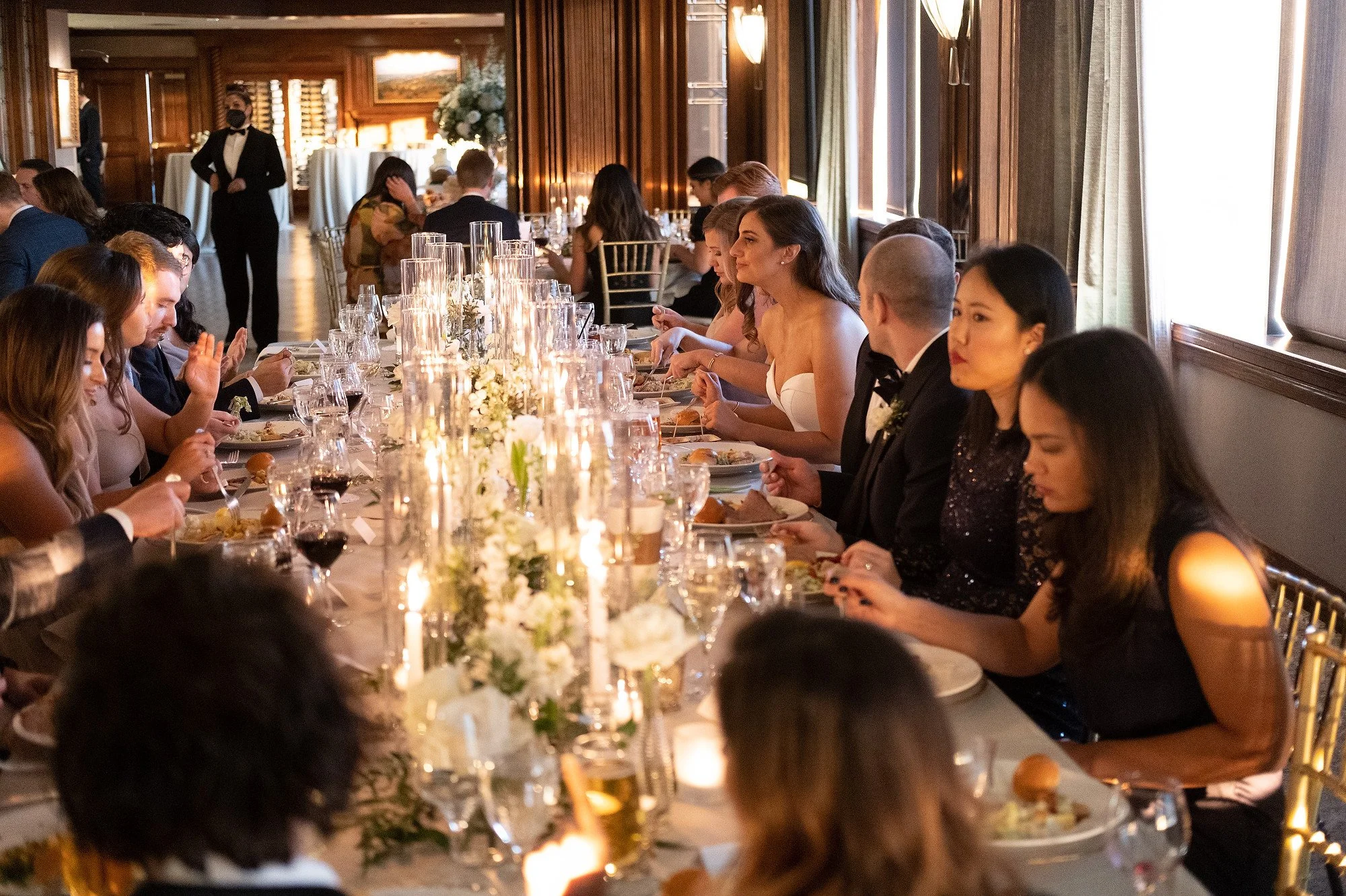People sitting at a long, elegant dinner table with candles and floral centerpieces during a formal event or wedding reception.