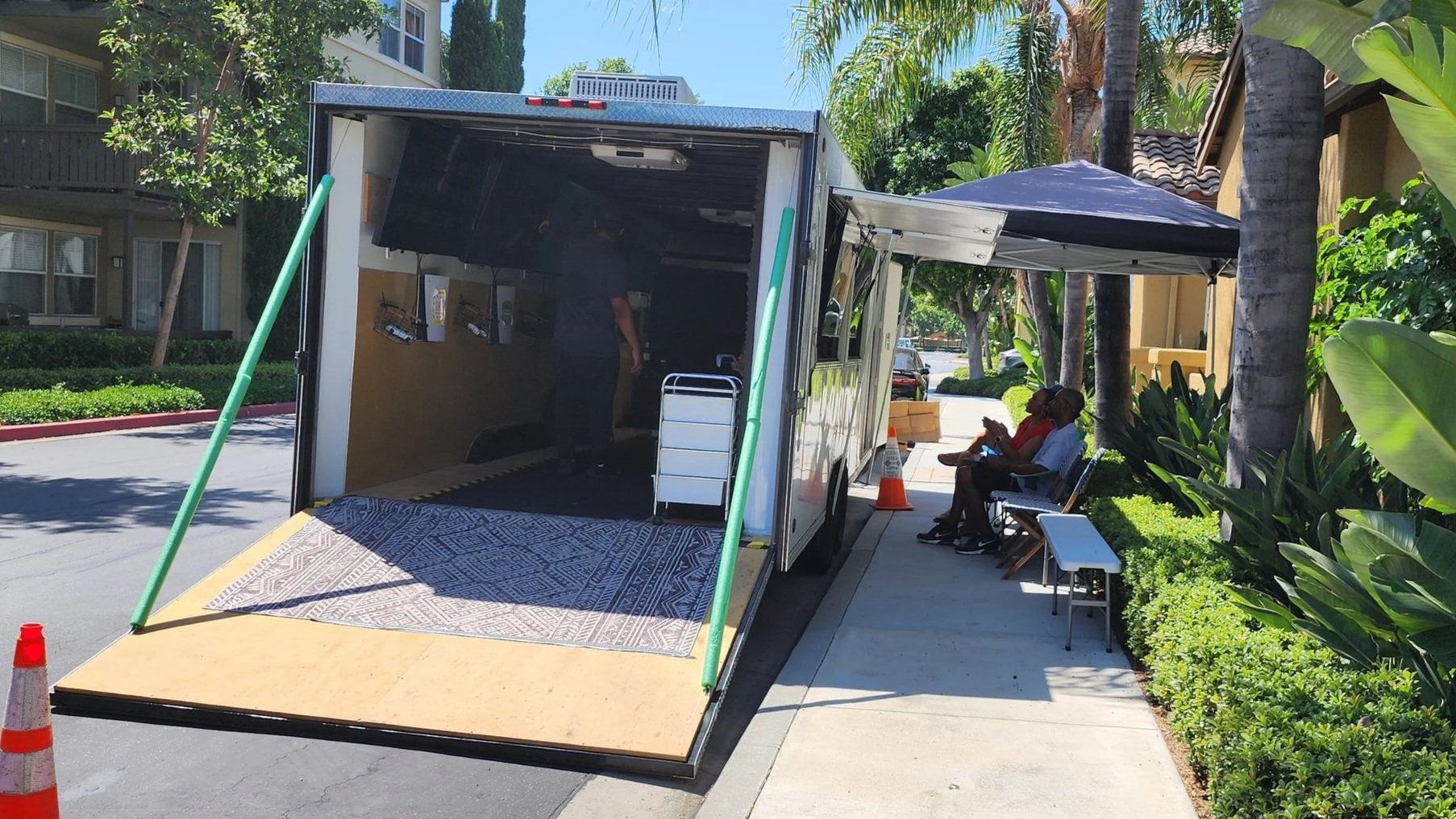 A video game truck is setup near an apartment complex.  There are 2 adults playing on the outside of the trailer.  The ramp of the trailer is down for easy access.