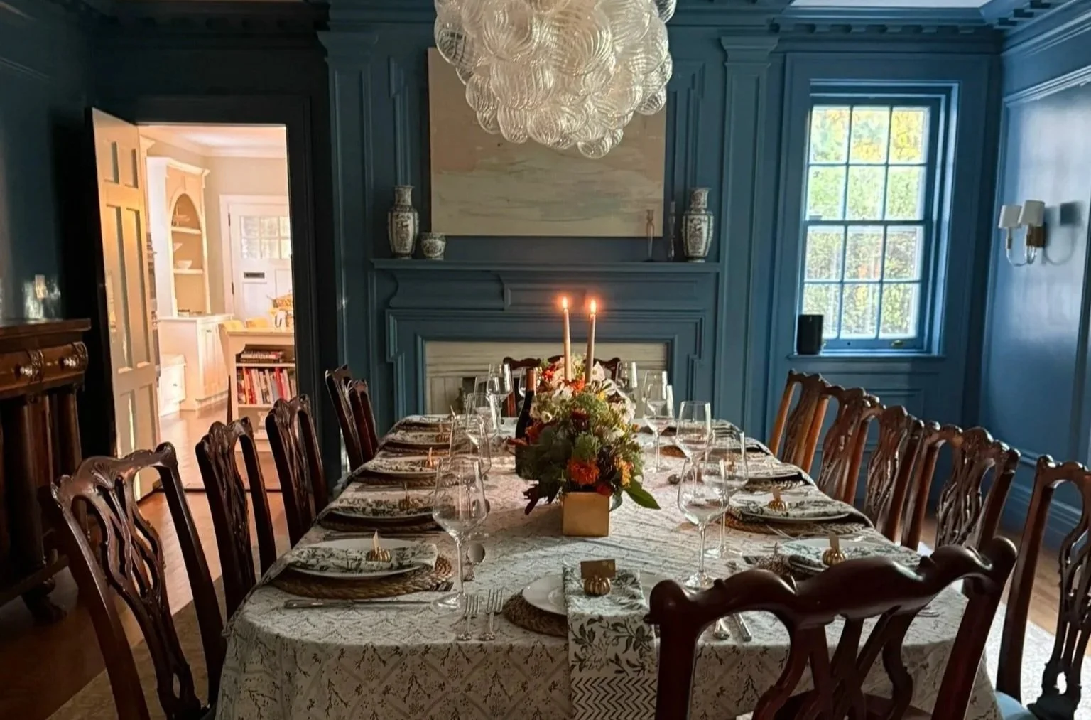 Elegant dining room in a historic home with a long table set with fine china, glassware, and a floral centerpiece with two candles. Blue walls with decorative molding, a large window, and a fireplace with vases on the mantel.