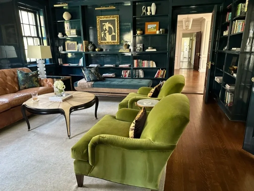 A historic home's library featuring lacquered blue walls, l green velvet armchairs, a camel leather sofa, a round coffee table, a built-in lacquered blue bookshelves, and a hallway in the background. Brass accents and classic lighting light the space
