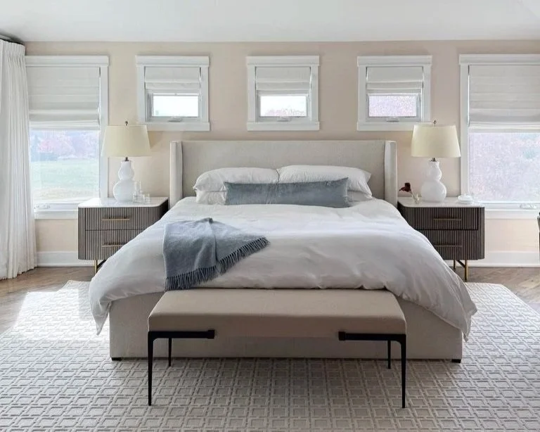 A tailored primary bedroom featuring a bright, neatly made bed with a white comforter, decorated with light blue velvet lumbar pillow and white pillows, surrounded by five windows with ivory linen shades, two nightstands with lamps, and a white rug.