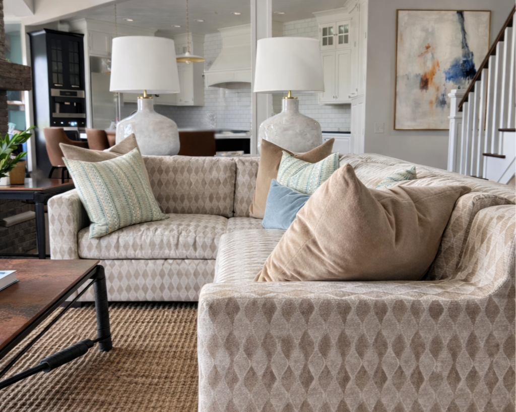 Coastal-inspired living room with custom tonal patterned sofa, decorative pillows, two large ceramic table lamps, a wooden coffee table, and artwork on the wall, with a view of the kitchen and staircase in the background.
