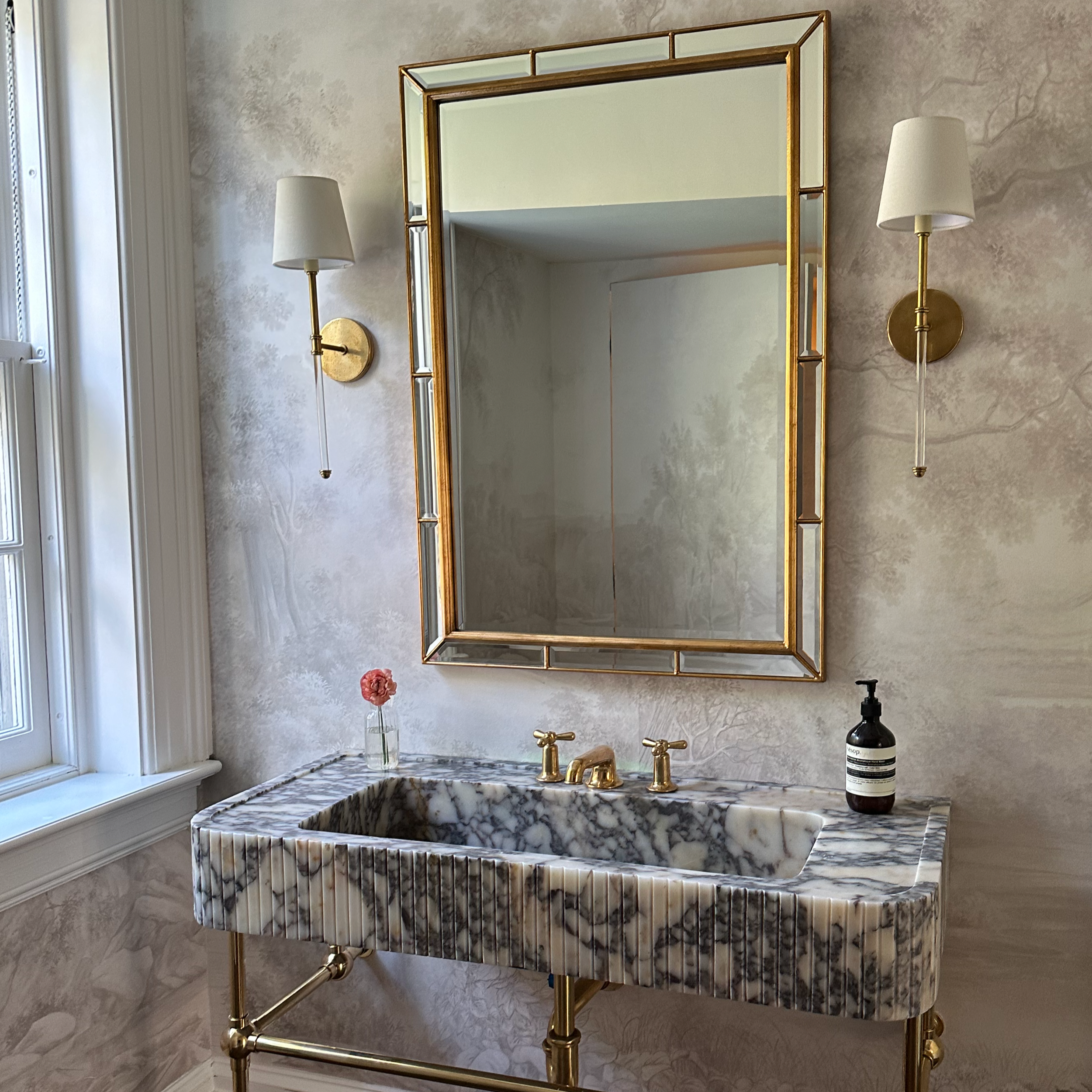 Bathroom with custom fluted calacatta viola marble sink, brass fixtures, gold-framed mirror, wall-mounted lamps, small pink flower in a glass bottle, and hand soap dispenser. Pastoral taupe mural on the wall.