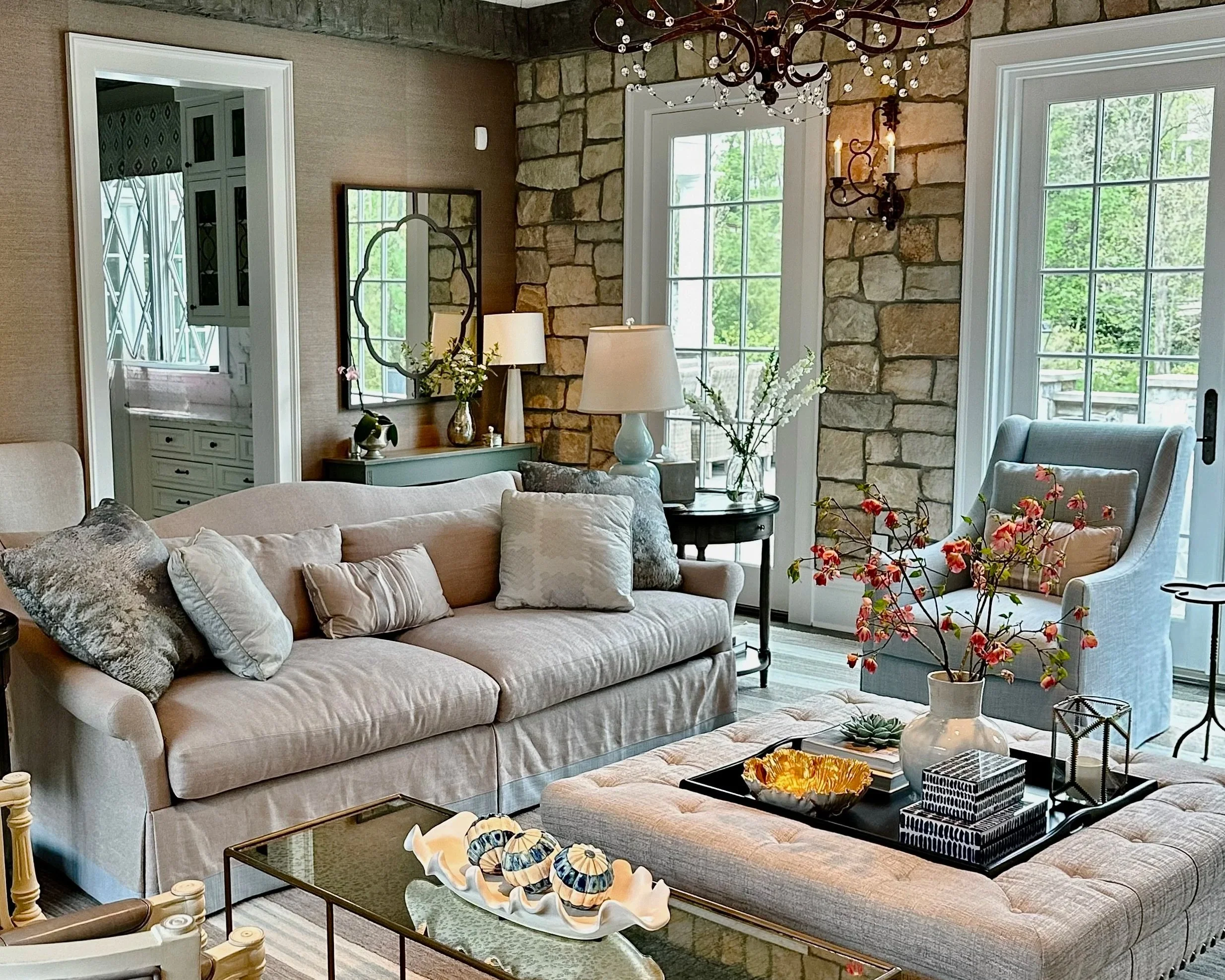 A cozy living room with a light-colored sofa adorned with decorative pillows, a tufted ottoman with a tray holding decorative boxes and a bowl, a light blue armchair with a pillow, a black side table with a large white lamp, a glass vase with pink an