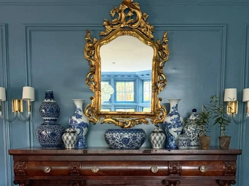 European-inspired collection of decorative blue and white porcelain vases and jars on a wooden sideboard, with a large ornate gold-framed mirror above, set against a blue wall with wall sconces.