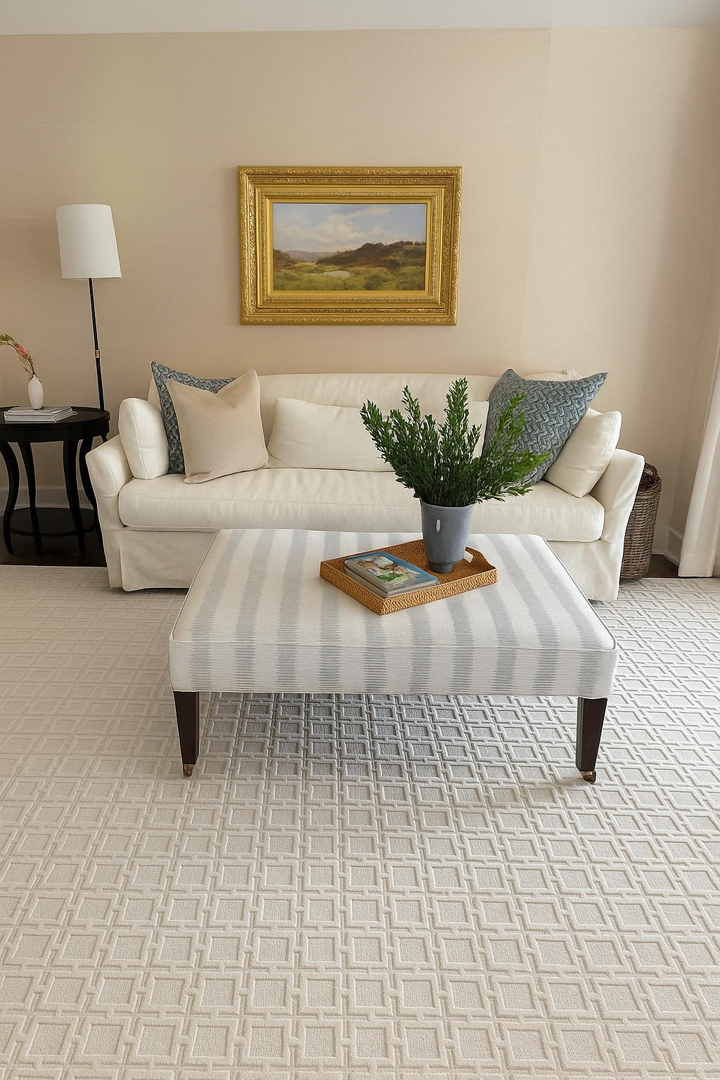 Living room with white sofa, framed landscape painting, side table with lamp and flowers, potted plant on striped ottoman, beige patterned carpet.