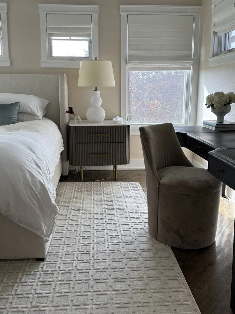 A cozy bedroom corner with a bed, nightstand, lamp, and a desk with chair and flowers.
