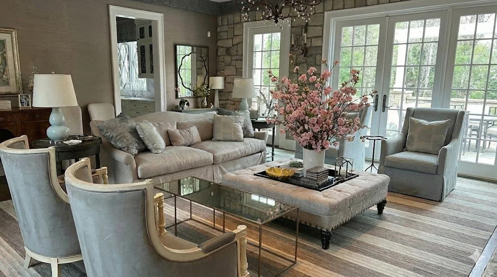 Living room with beige sofa, light-colored armchair, glass coffee table with floral arrangement, and large windows with glass doors leading outside patio.
