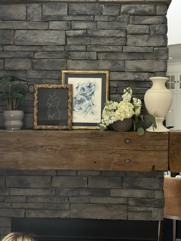 Decorative arrangement on a wooden mantel with picture frames, a white vase, a bouquet of white flowers, and a potted plant against a gray brick wall.