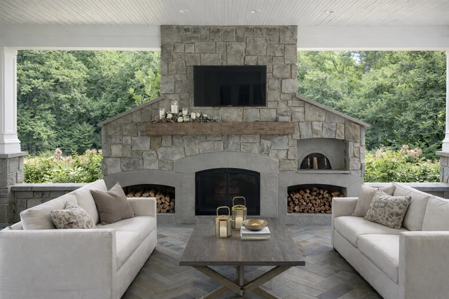 Outdoor living area with two white sofas, a wooden coffee table with lanterns and a decorative bowl, and a stone fireplace with a wall-mounted TV, surrounded by lush green trees.