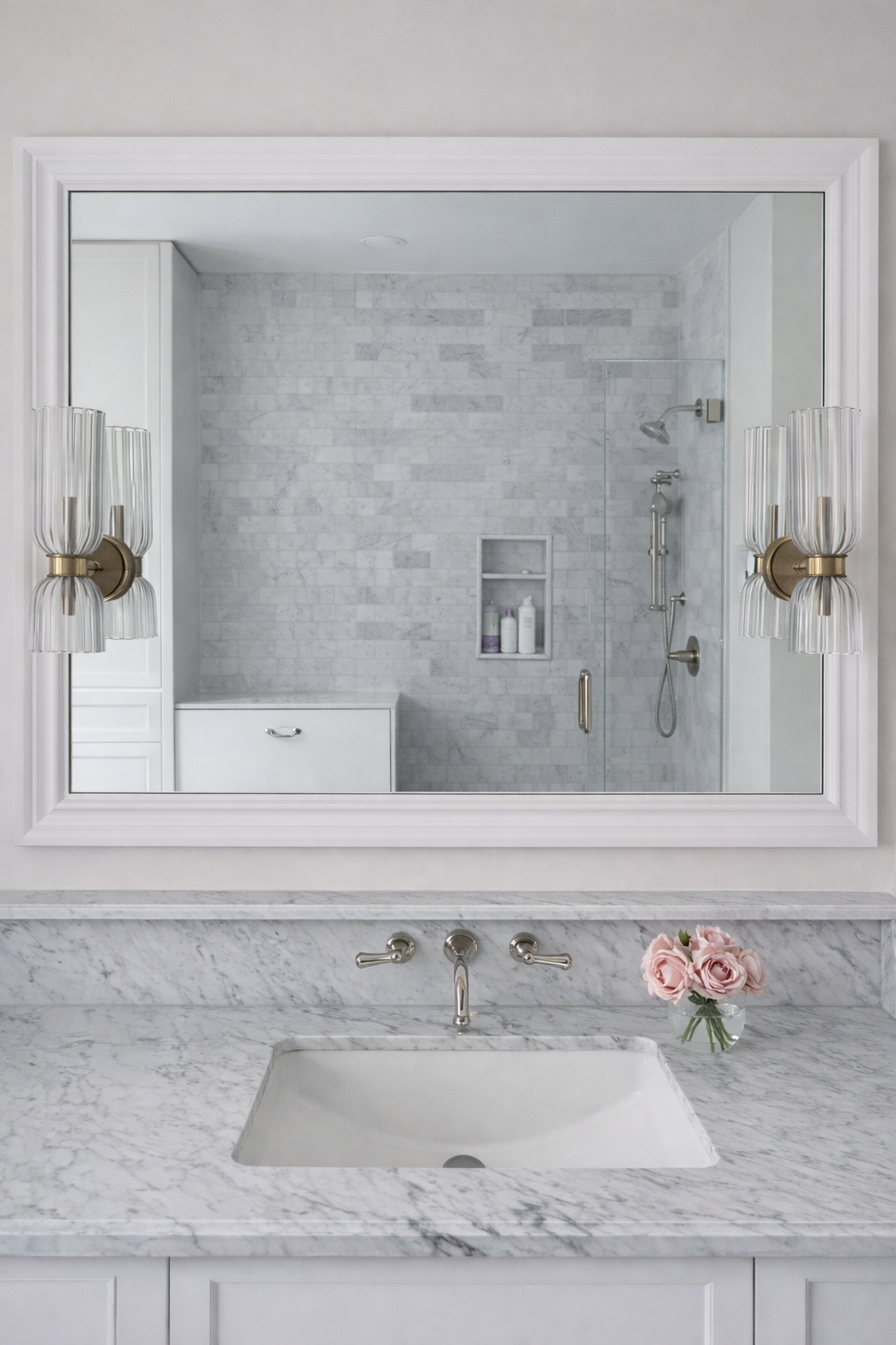 Elegant carrara marble bathroom featuring vanity with marble ledge, polished nickel fixtures, and a mirror reflecting a marble shower with niche and built-in white cabinetry.