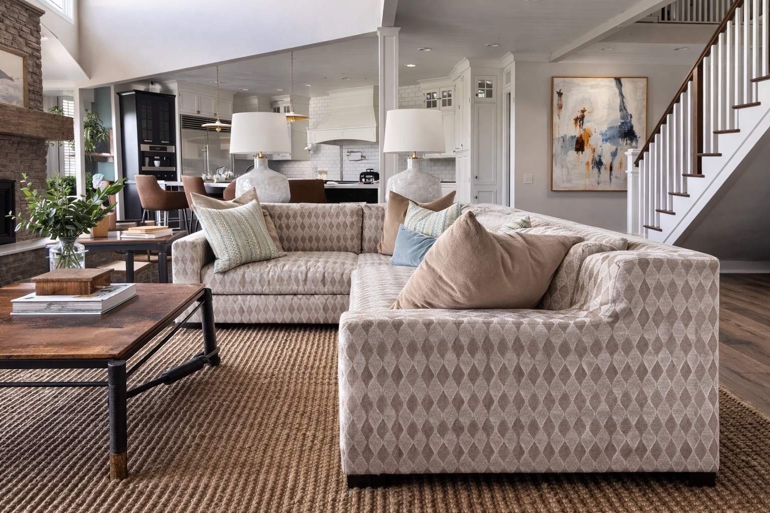 Living room with a beige sectional sofa, decorative pillows, coffee table, large lamps, and open kitchen area in the background.