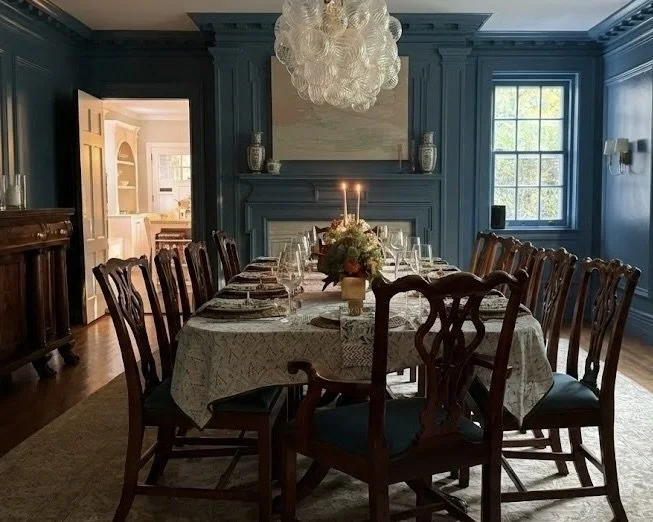 A formal dining room with a long table set for dinner, decorated with a floral centerpiece and candles. The room has dark blue walls, a large window, a chandelier hanging from the ceiling, and a fireplace with decorative vases on the mantel.