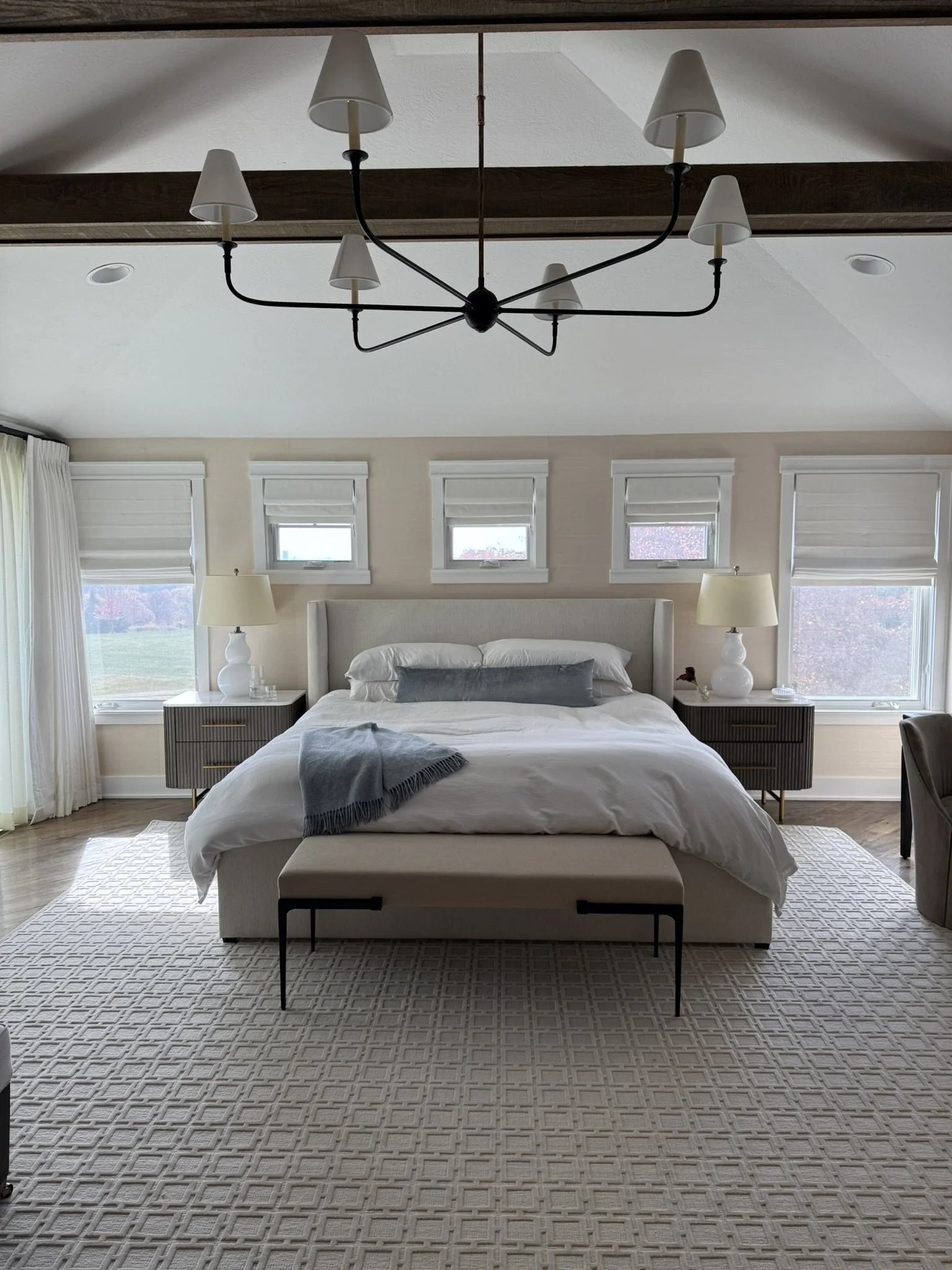 A cozy bedroom with a large bed, white bedding, a gray throw blanket, and multiple white pillows. Flanking the bed are two matching nightstands with lamps. There are four small windows above the headboard and a patterned area rug on the wooden floor.