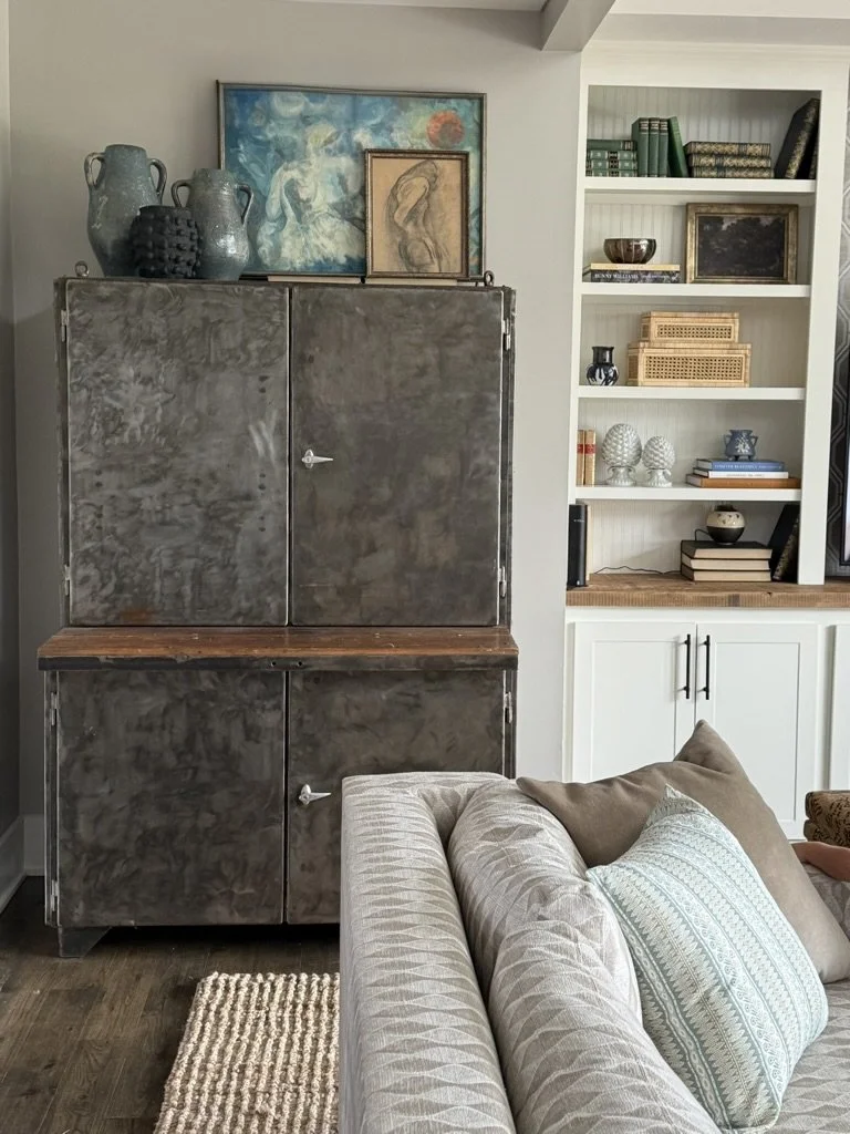 Living room with a metal storage cabinet, artwork, and decorative vases on top; white bookshelf with books and decorative objects; beige couch with patterned pillows in foreground.