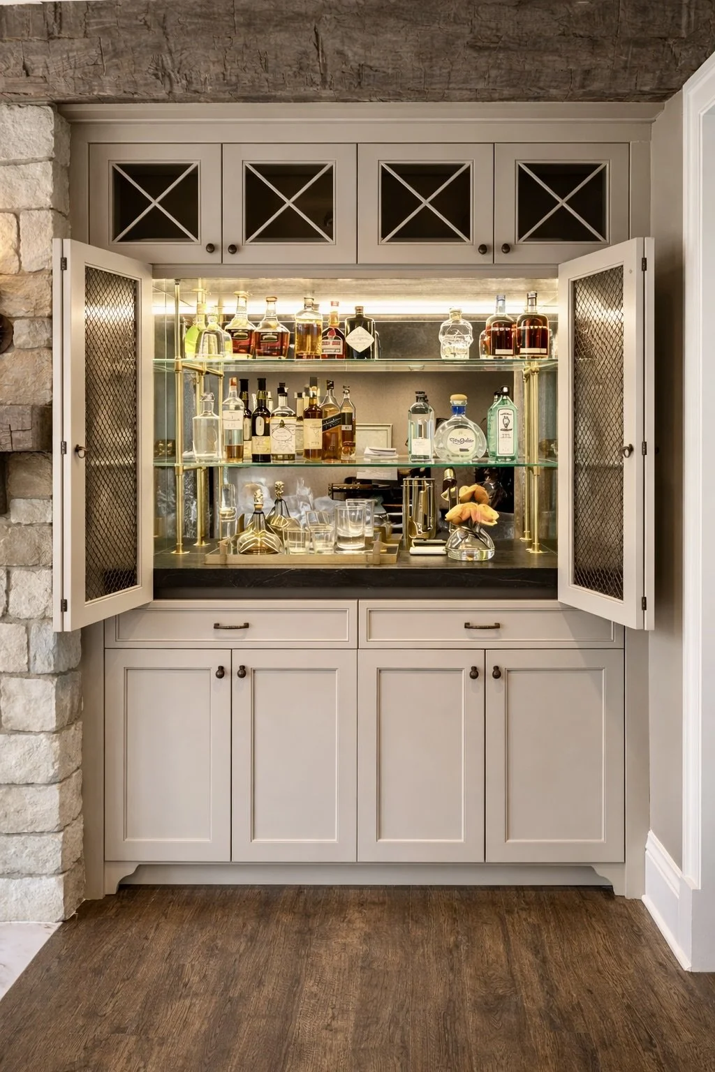 A custom built-in bar featuring brass tubular poles, glass shelves and antique mirror backsplash. The shelves are filled with assorted bottles of alcohol, glasses, and accessories, with a rustic stone and wood interior.
