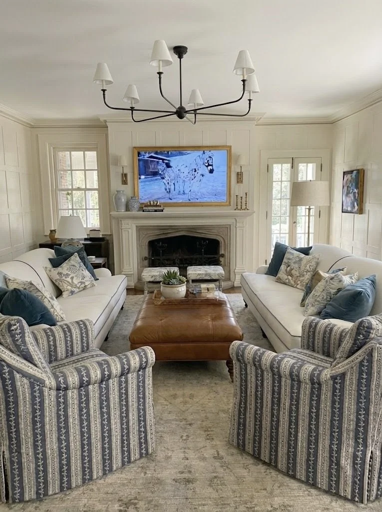Historic home's living room with two white sofas, two striped armchairs, a leather ottoman, a fireplace, a flat-screen TV, multiple decorative pillows, side tables with lamps, and a chandelier on the ceiling.