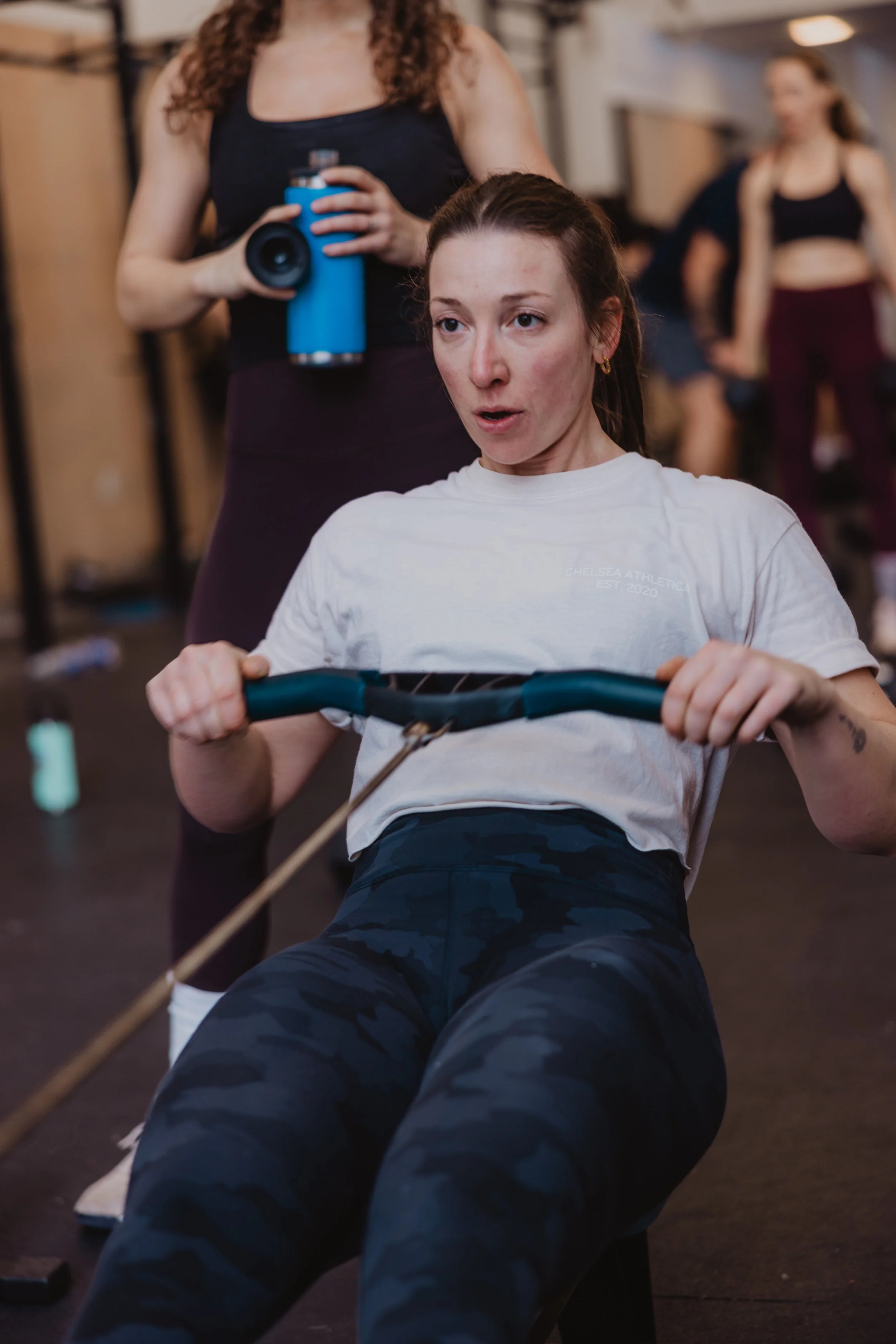 A woman in a white T-shirt and camouflage leggings is doing a rowing exercise on a machine in a gym, with a partner standing behind her holding a water bottle.