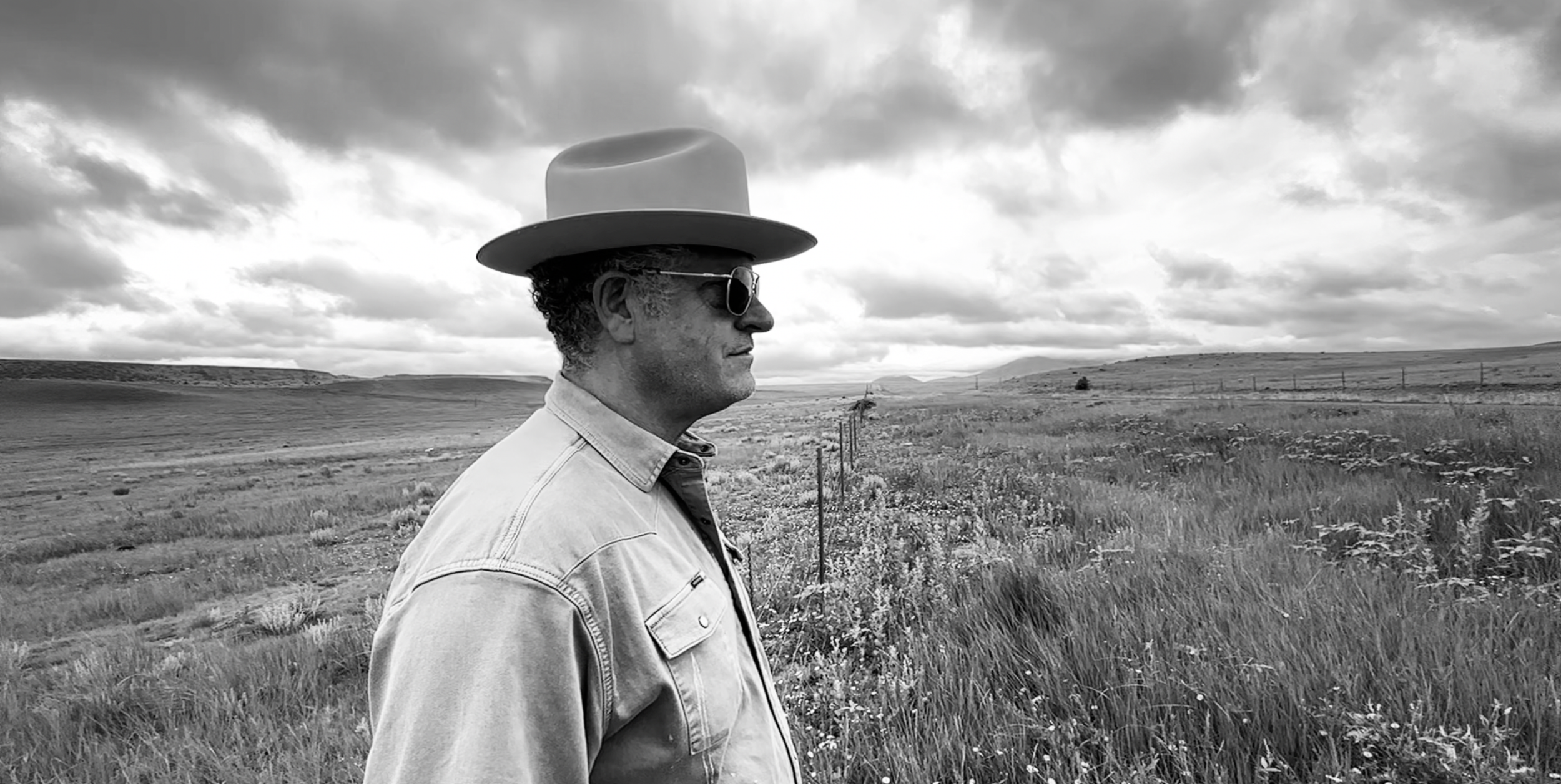 A man in sunglasses and a wide-brimmed hat stands in profile in a rural landscape with fields, a fence, and hills in the background under a cloudy sky.