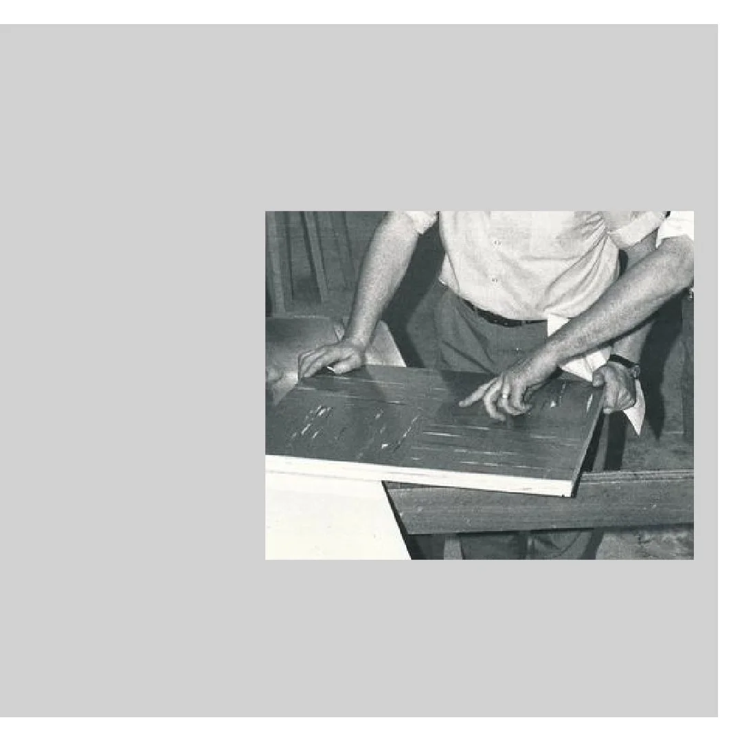 A person wearing a light-colored shirt appears to be working on a wooden surface, possibly sanding or smoothing it, in a workshop or room with wooden furniture.