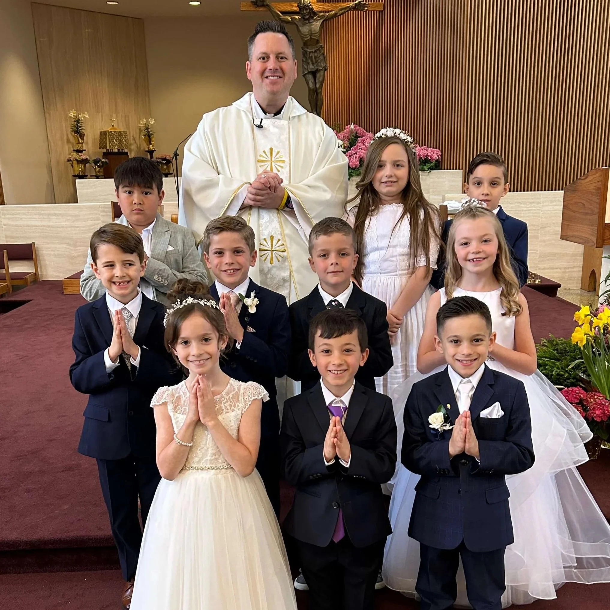 Young children in Holy Communion outfits, smiling with prayer hands, standing with young priest
