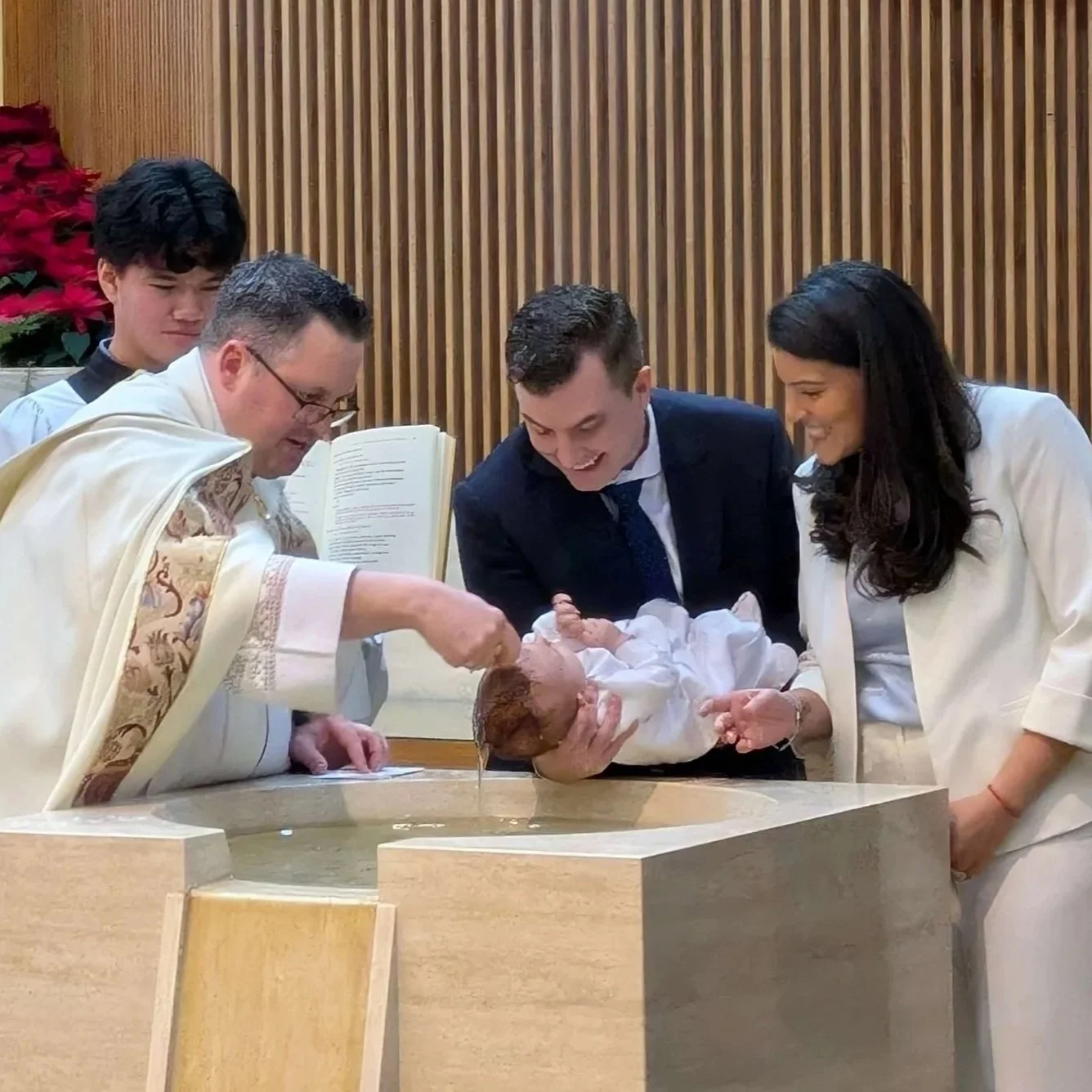 Mother and father holding and smiling at baby held over baptismal fountain as Father Paul gently pours water on the baby's head