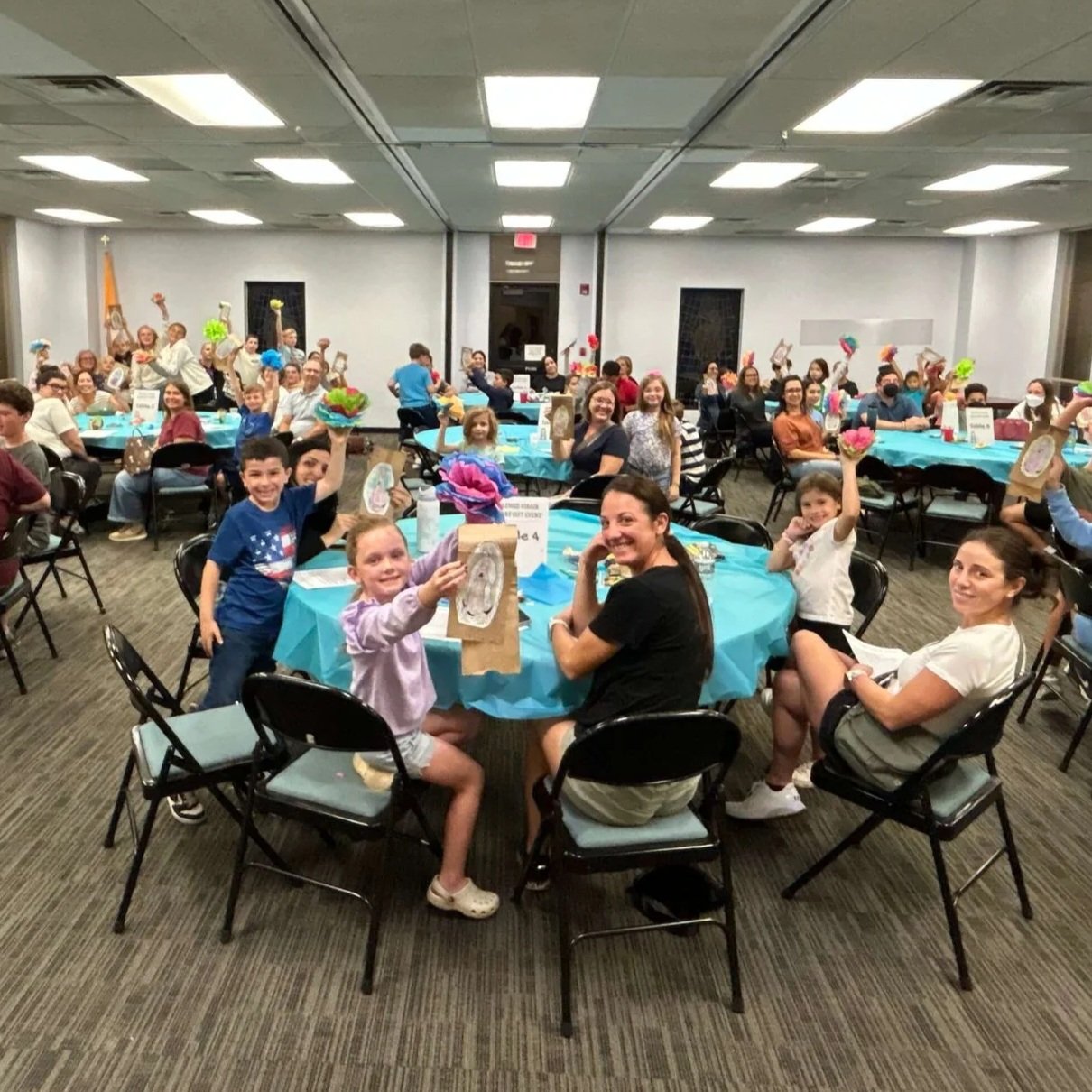 Families smiling around tables in a room, with children holding up a craft