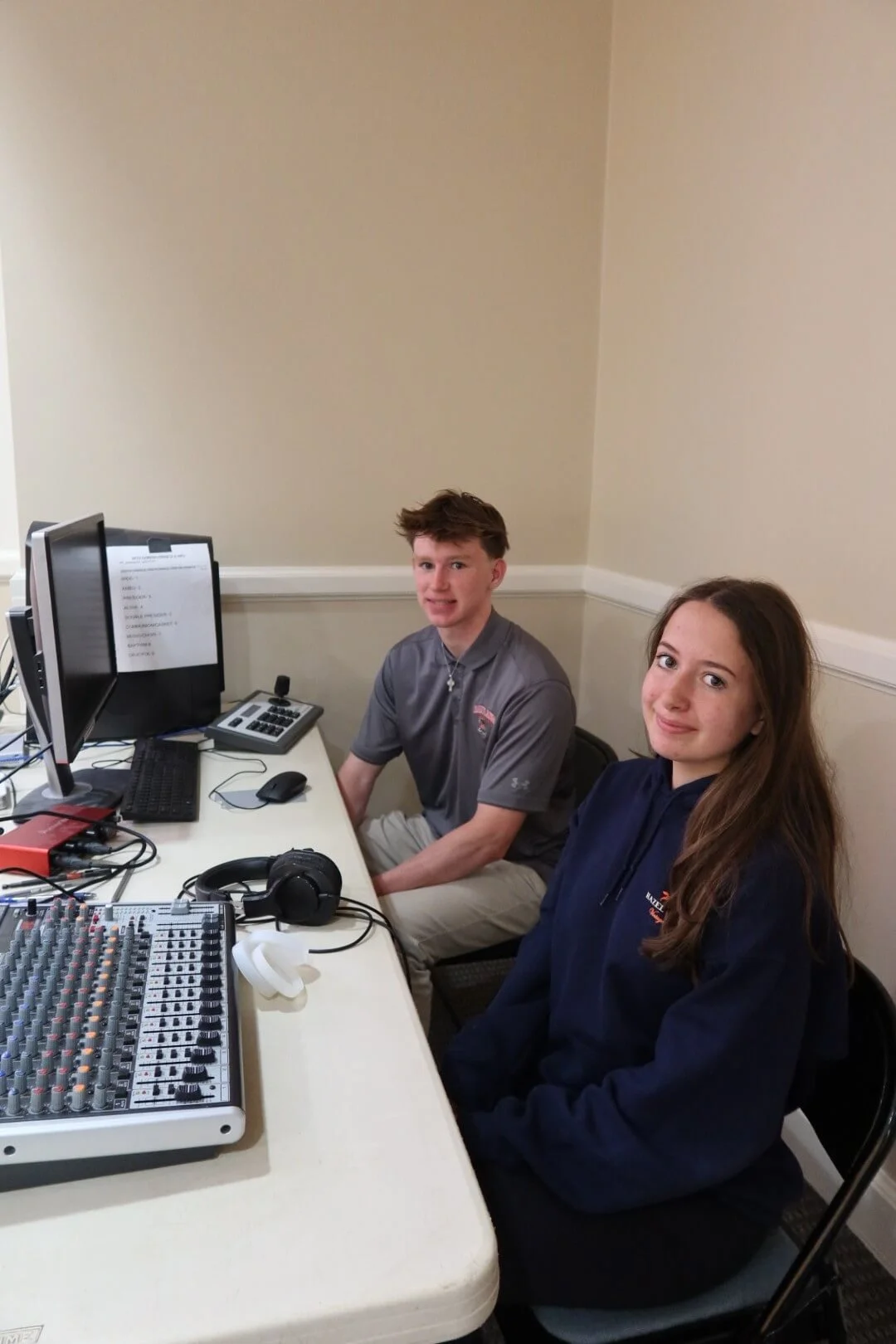 Teen boy and girl sitting with computer monitor, headphones and equipment, smiling.