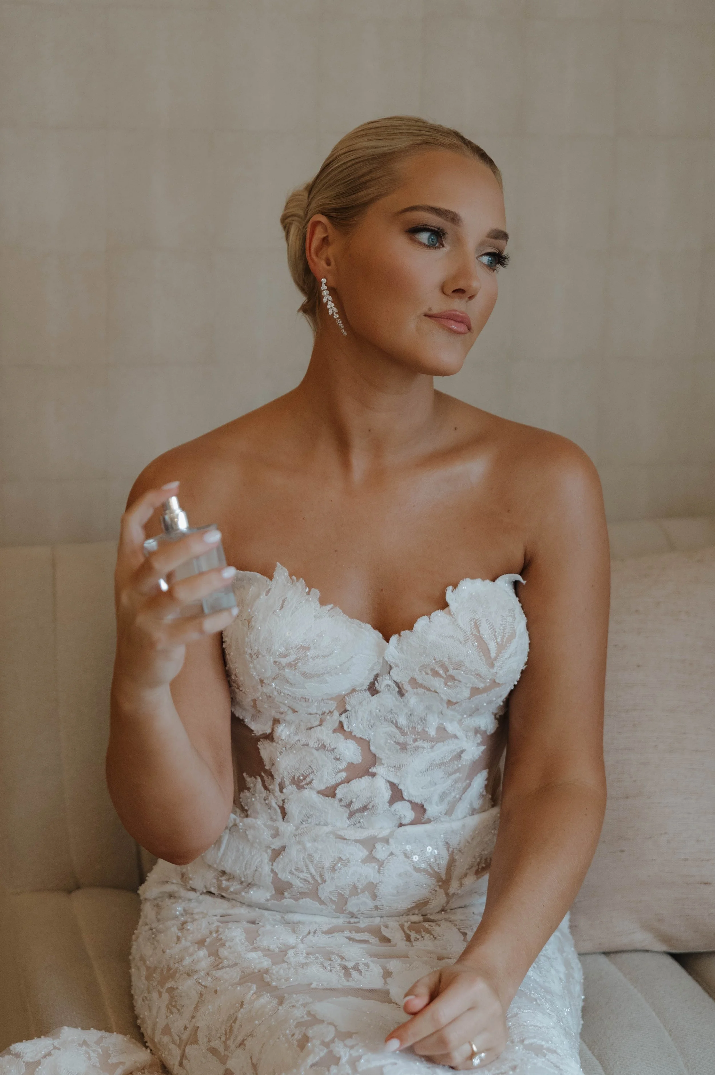 A bride putting on earrings, wearing a white lace wedding dress with her eyes closed.