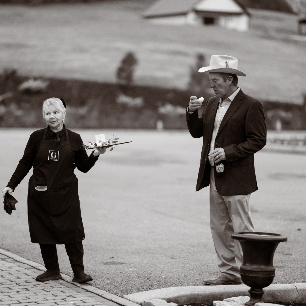 A man in a suit and cowboy hat eating an ice cream cone while standing on the sidewalk, and a woman in a black apron holding a tray with food or drinks standing nearby outdoors.