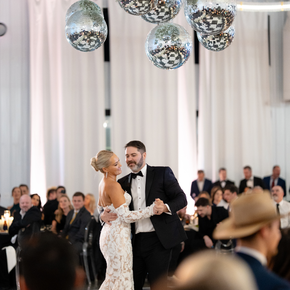 Couple dancing at a wedding reception with guests seated around them and disco balls hanging from the ceiling.