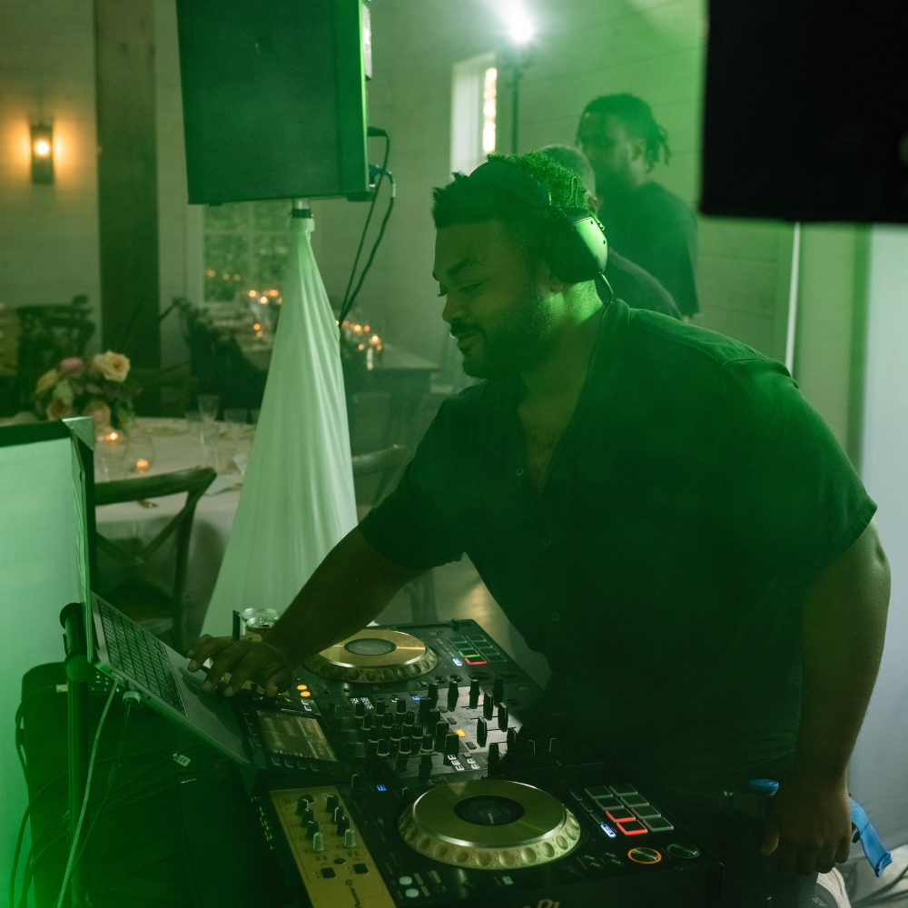 A DJ wearing headphones mixing music on a DJ controller at an indoor event with round tables and floral centerpieces, illuminated with green lighting.