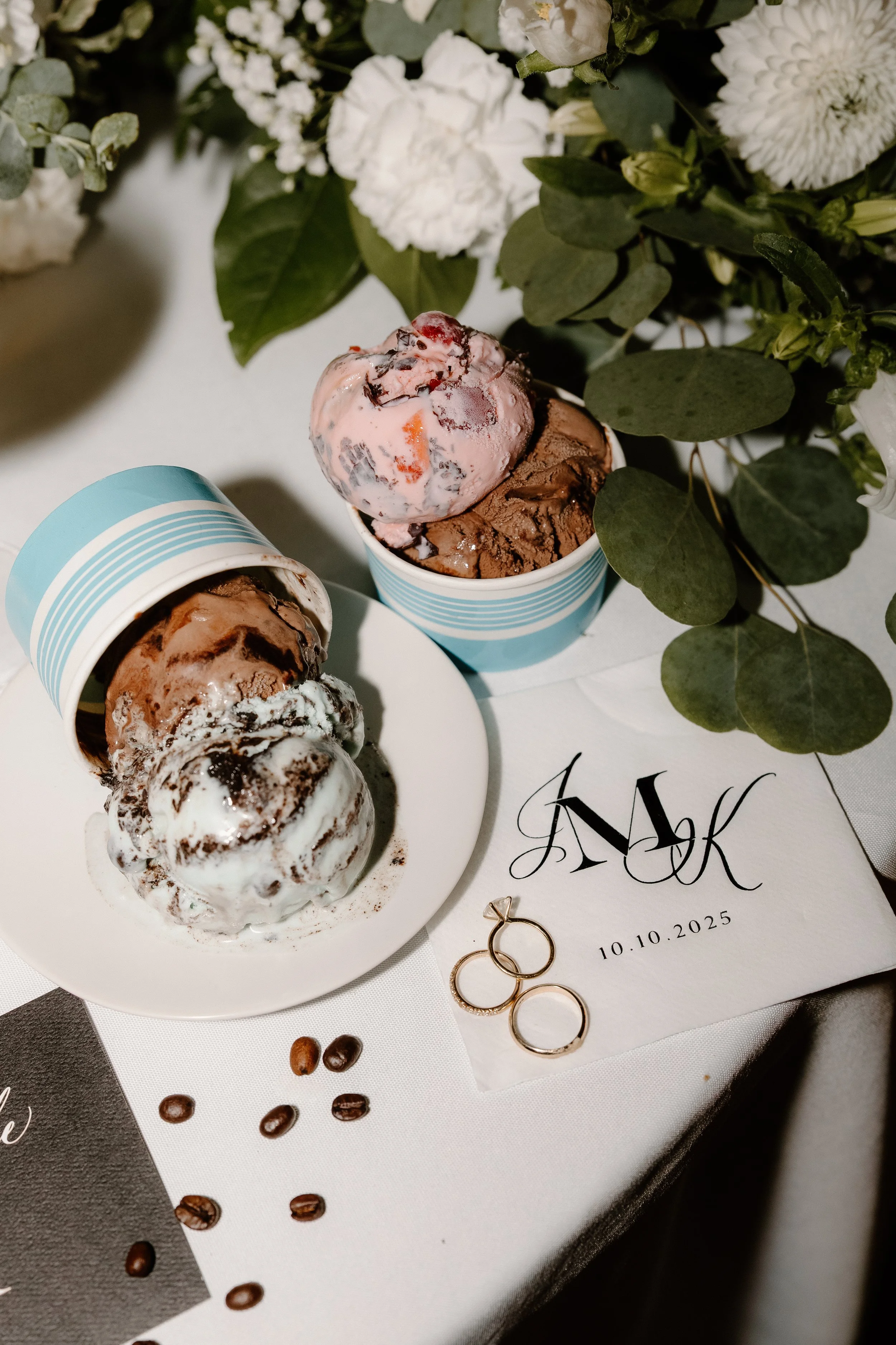 Three cups of assorted ice cream on a white table, with a bouquet of flowers and wedding rings nearby.