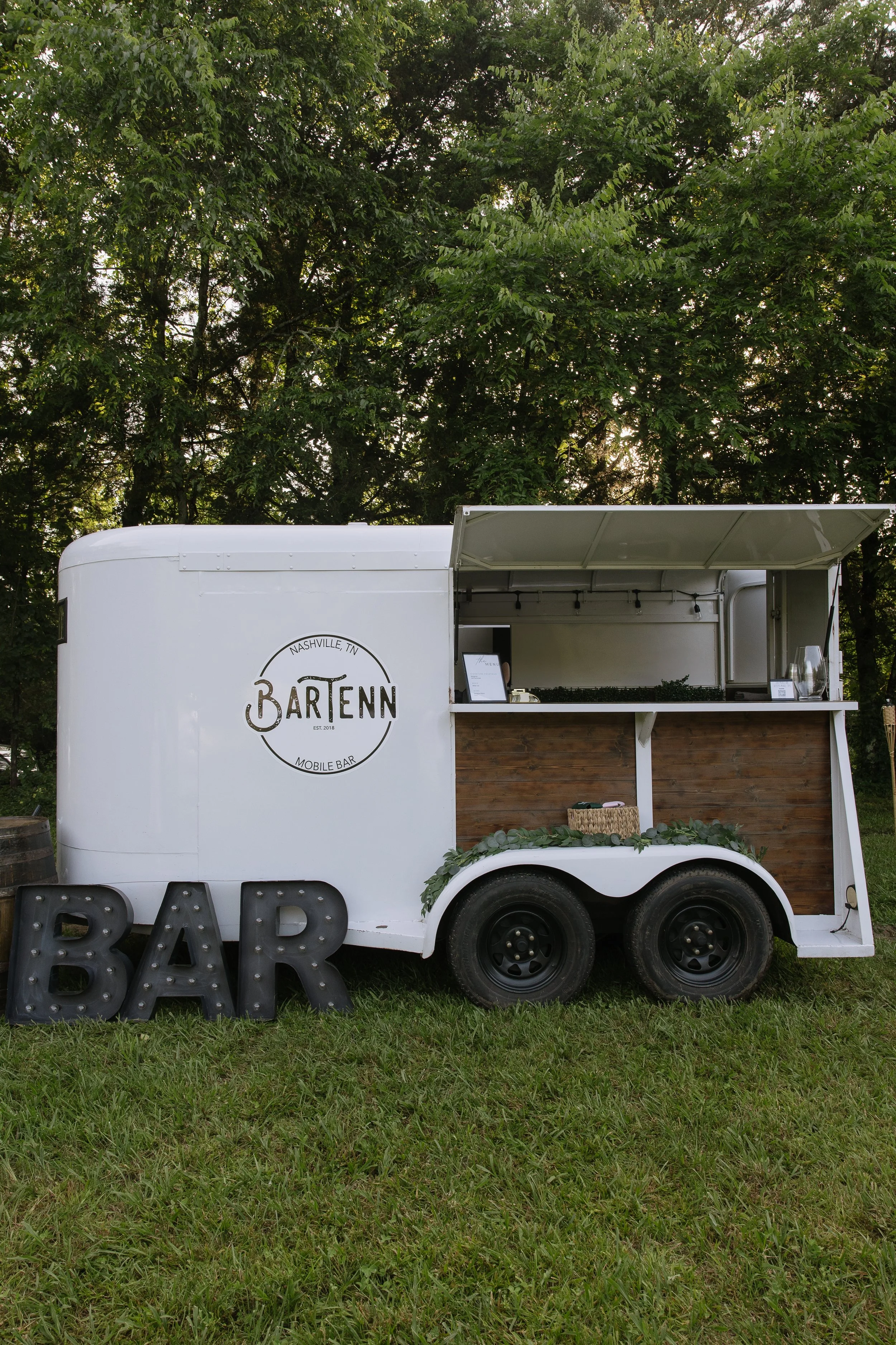 A white mobile bar trailer with the logo "BARTENN Nashville, TN Mobile Bar" and a large black "BAR" sign in front. The trailer is set outdoors on grass, with a backdrop of green trees.