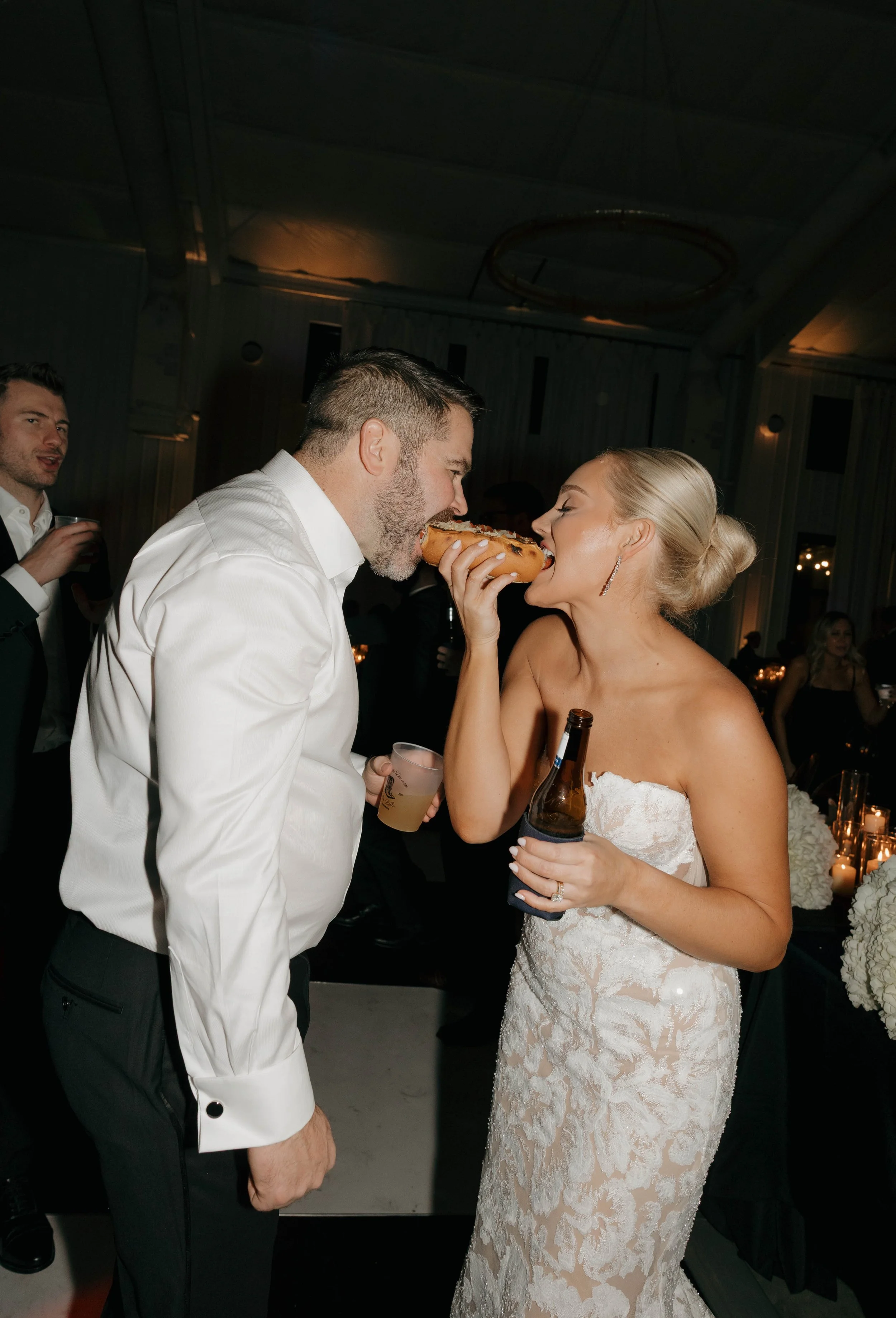 A bride and groom share a moment at their wedding reception, with the bride holding a beer bottle and the groom holding a drink, while the bride gets ready to take a bite of a hot dog being offered by the groom.