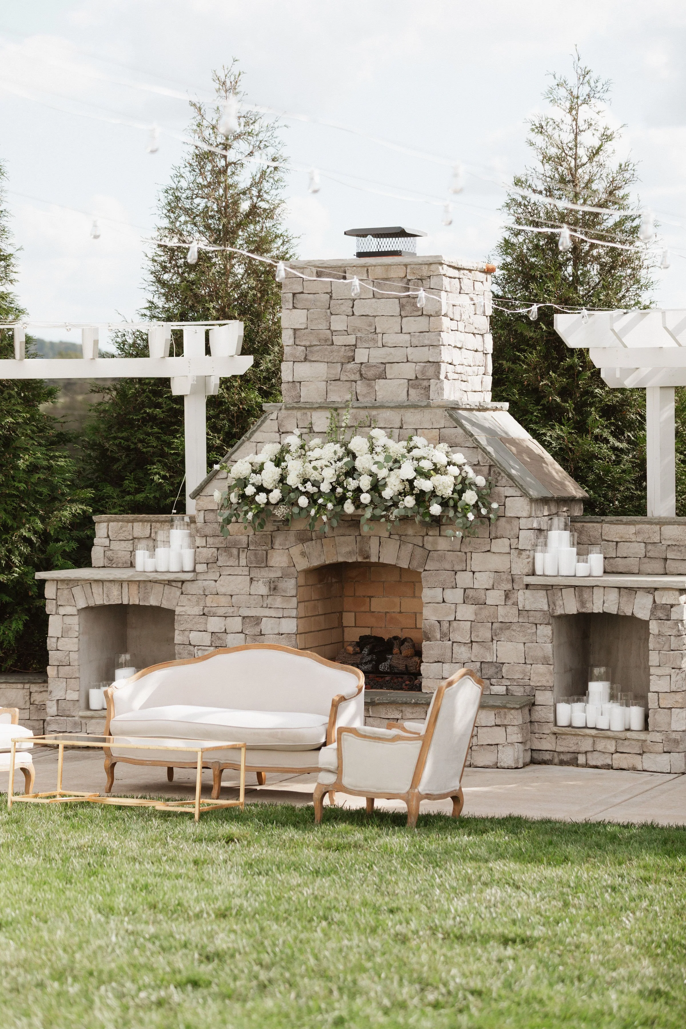Outdoor patio area with a stone fireplace decorated with white flowers and candles, surrounded by vintage-style furniture and string lights, set against a backdrop of green trees.