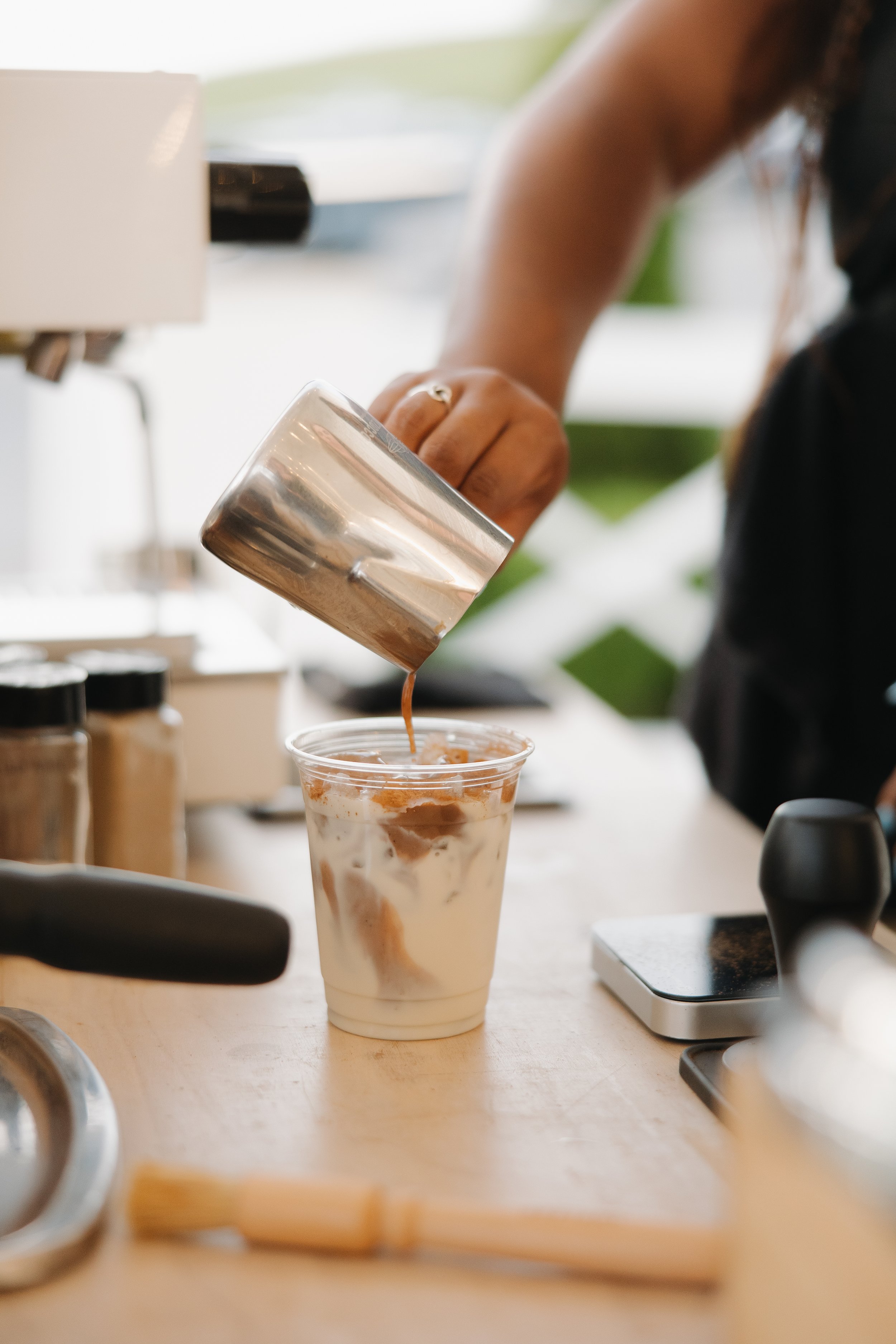Person pouring coffee into a plastic cup, with coffee-making tools on a wooden table.