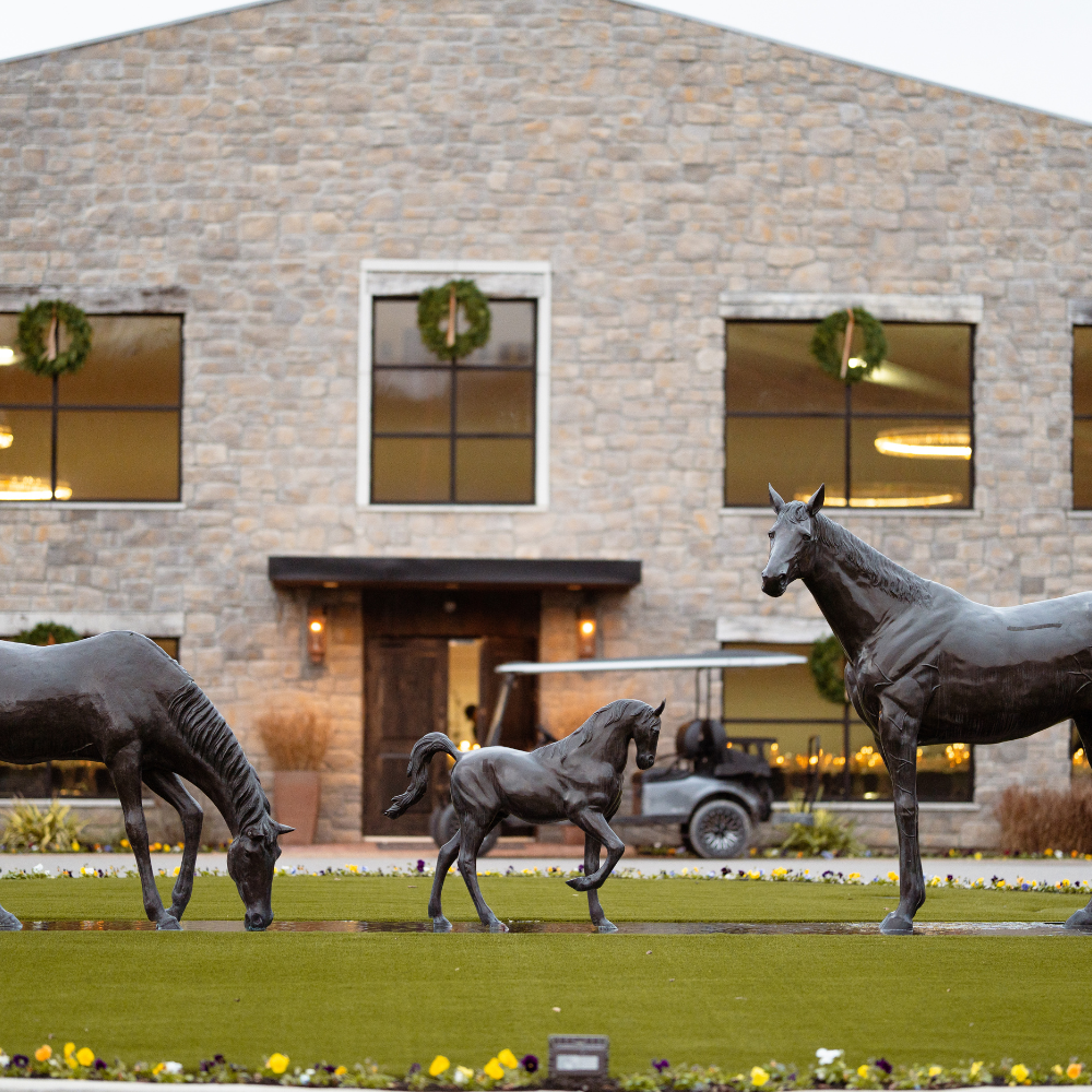 Bronze statues of three horses, including a foal, on a lawn in front of a stone building with large windows decorated with wreaths, and a golf cart parked nearby.